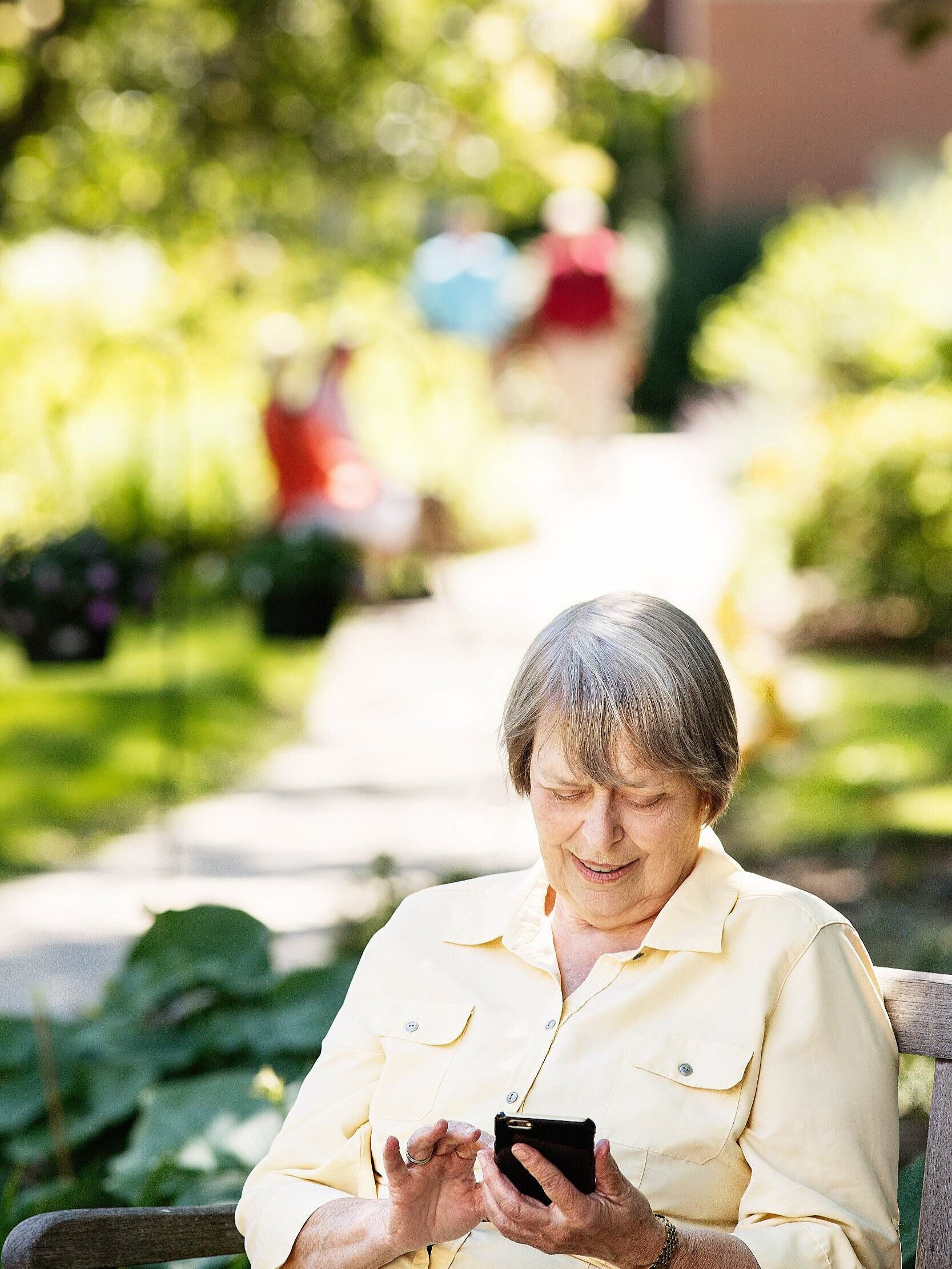 An elderly woman sitting on a park bench, smiling while looking at her smartphone with a garden and blurred people in the background.