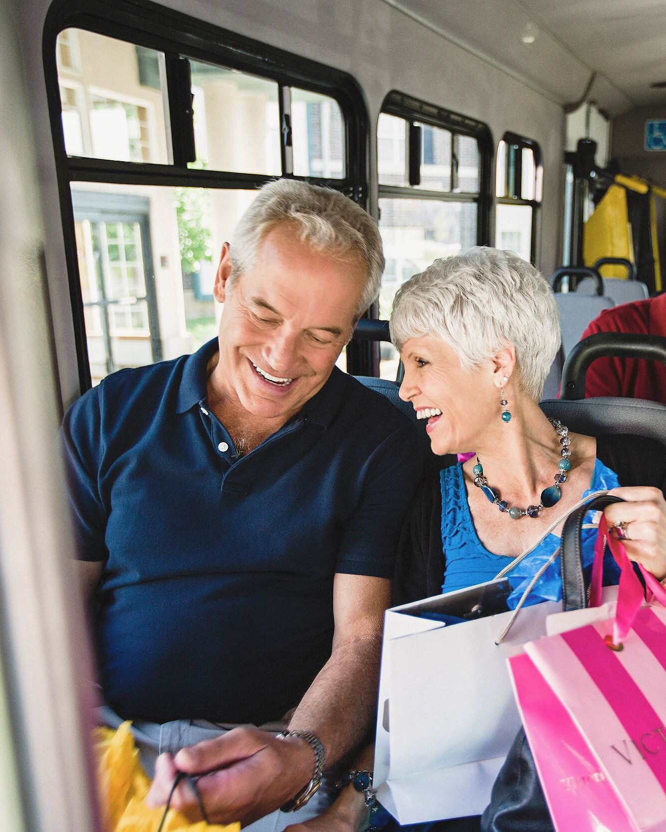 A smiling elderly man and woman sitting together on a bus, exchanging happy looks while surrounded by shopping bags.