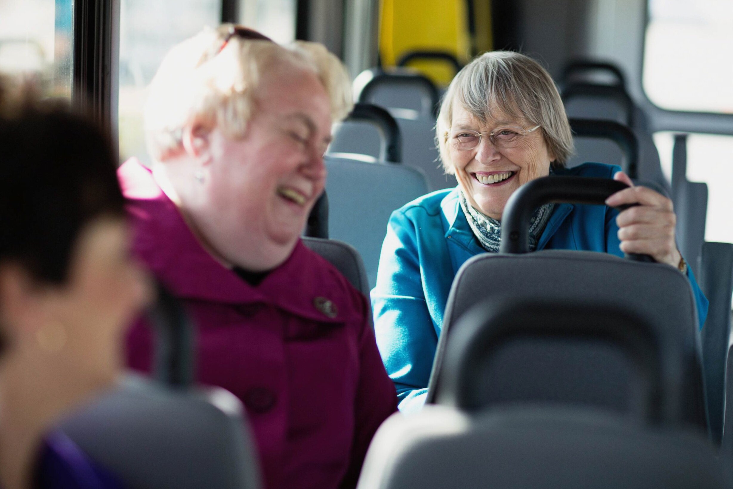 Two women laughing and smiling on a bus, one older woman with glasses and a blue jacket, the other younger woman with blonde hair and a purple jacket, sitting in seats with gray upholstery and large windows