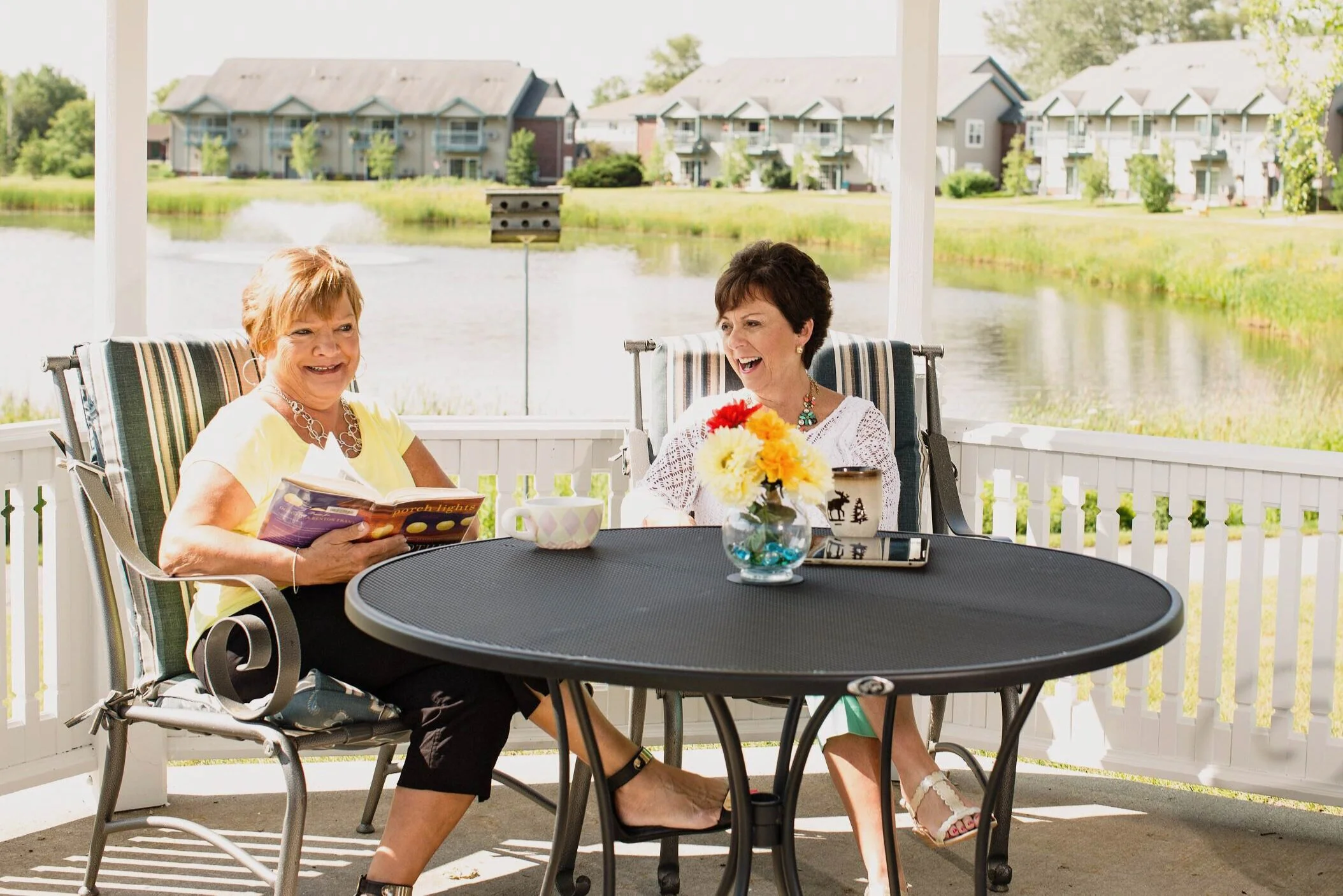 Two women sitting at a round table on a screened porch, laughing and enjoying each other's company with a lake and houses in the background.