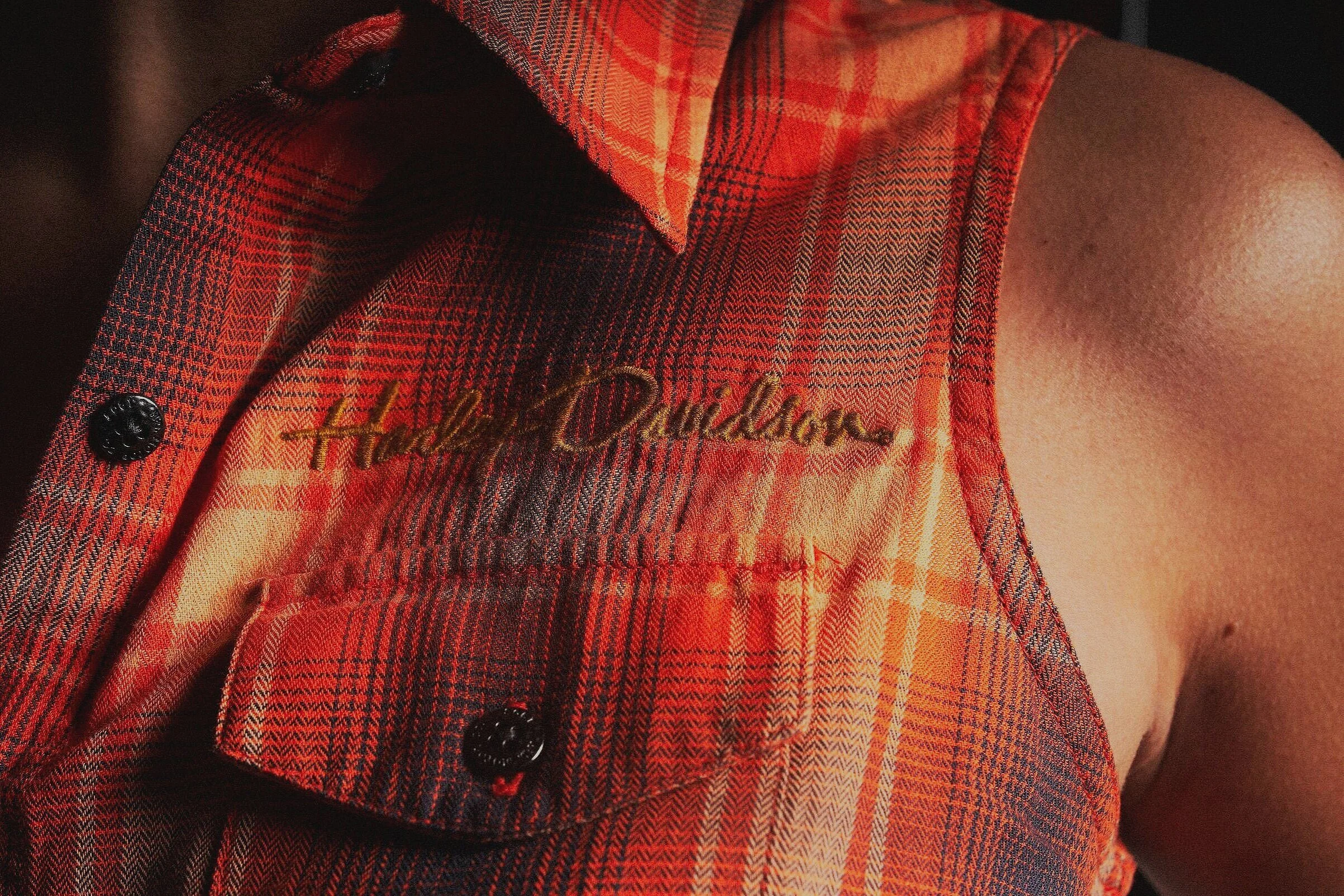 Close-up of a person's sleeveless plaid shirt in red and orange tones with black buttons, embroidered with the words 'Hello Darling' on the chest pocket.