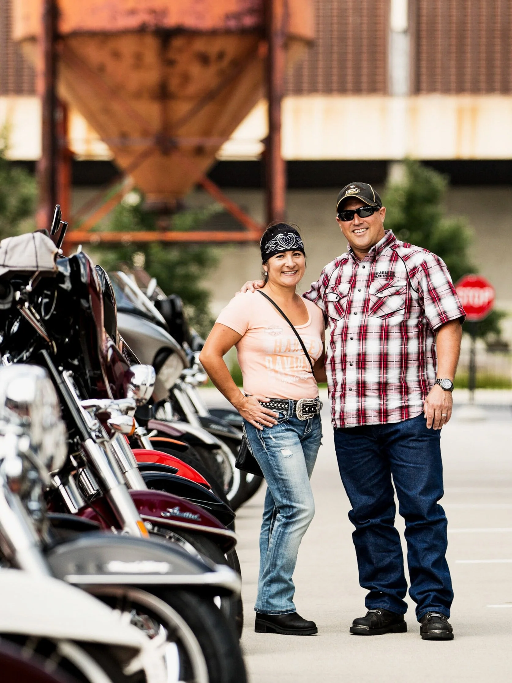 Two smiling people, a woman and a man, standing next to motorcycles in a parking lot. The woman has dark hair, is wearing a bandana, a peach T-shirt, ripped jeans, and black boots. The man has short hair, is wearing sunglasses, a plaid shirt, and dar