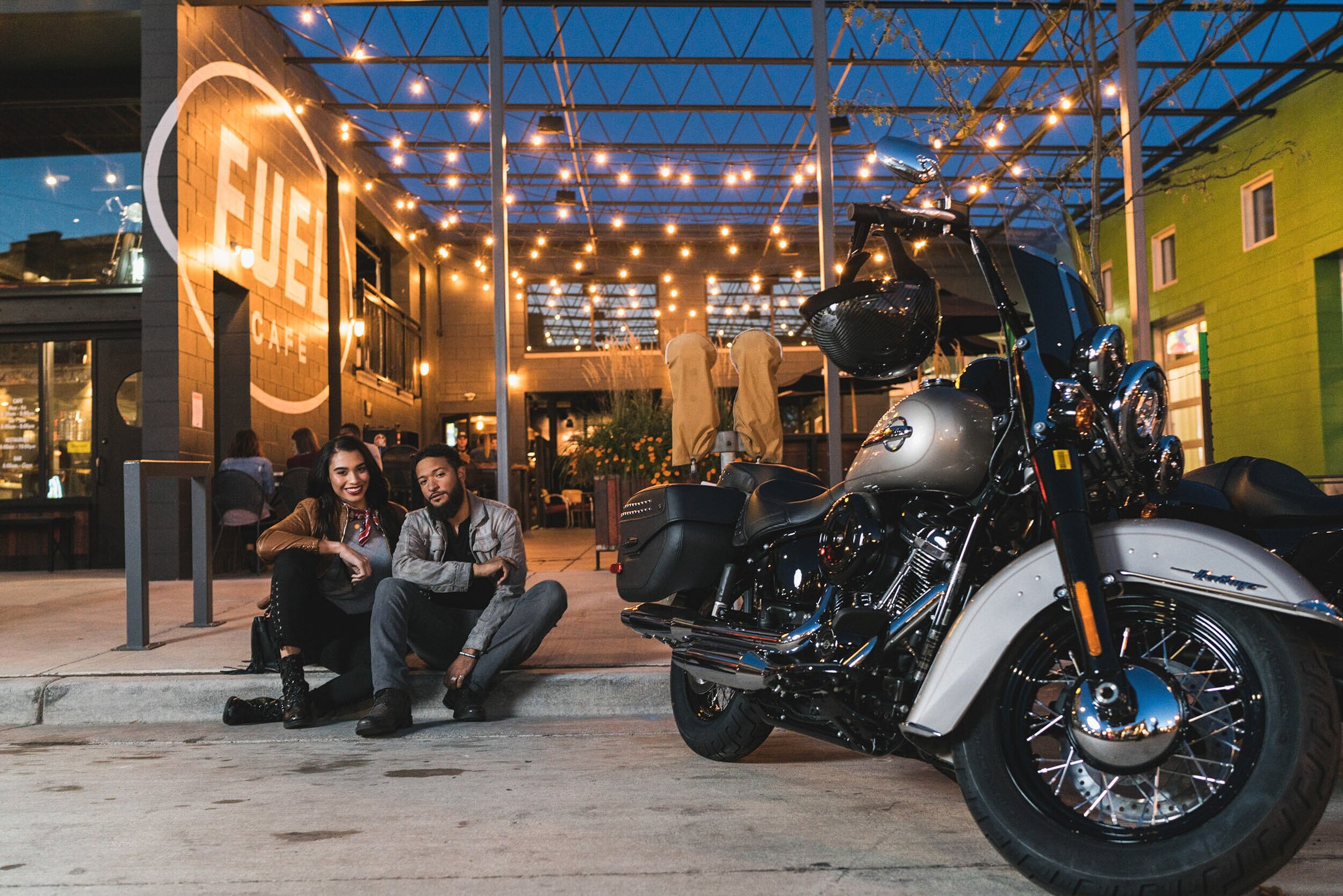 Two people sitting on the curb outside a cafe with a large motorcycle parked nearby during the evening. The cafe has string lights hanging from a metal framework overhead, and there's a large sign that reads "FUEL CAFE" on the wall.