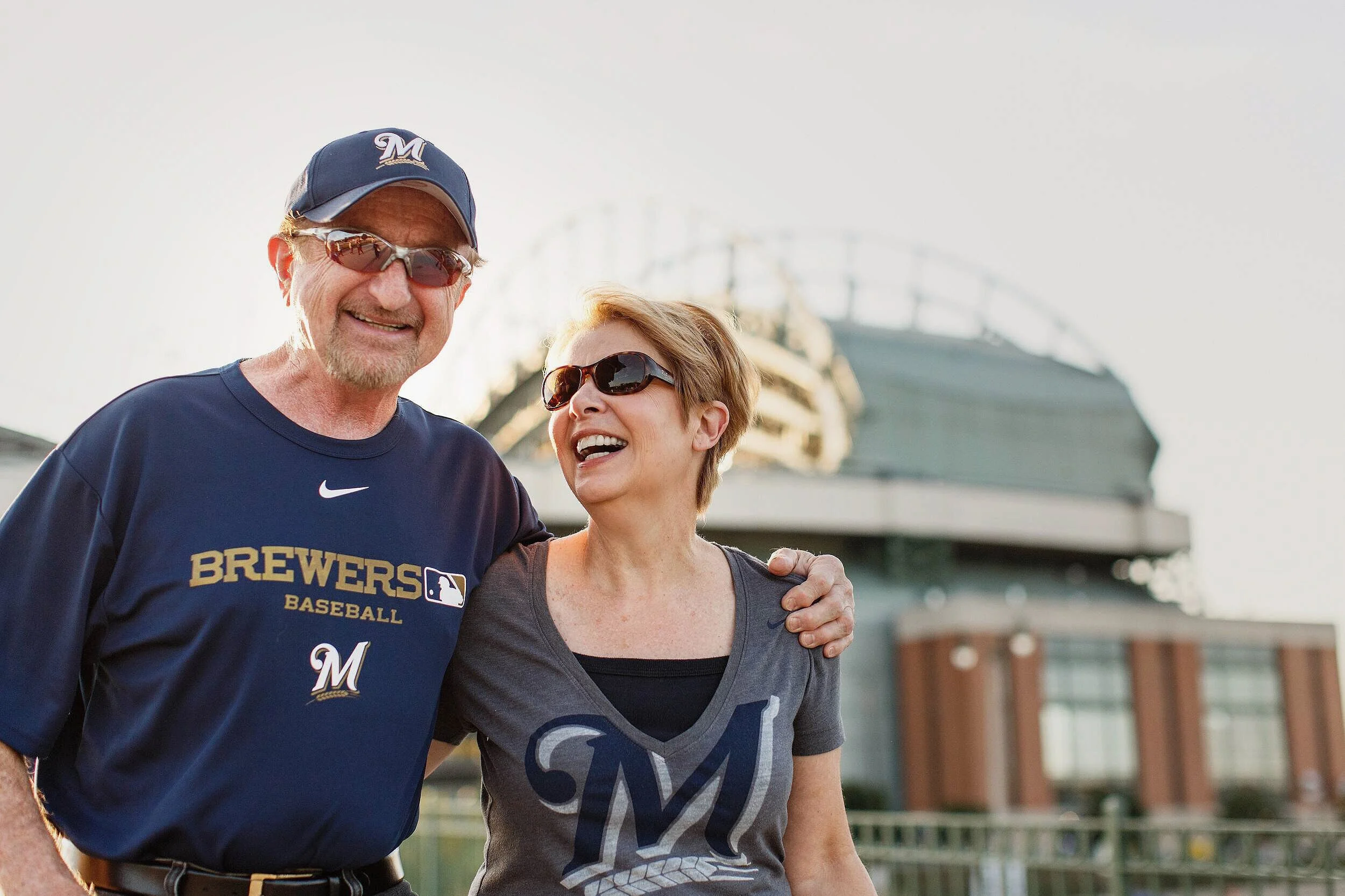 A smiling man and woman wearing Milwaukee Brewers apparel and sunglasses, standing outdoors in front of a stadium, with the woman’s arm around the man’s shoulder.