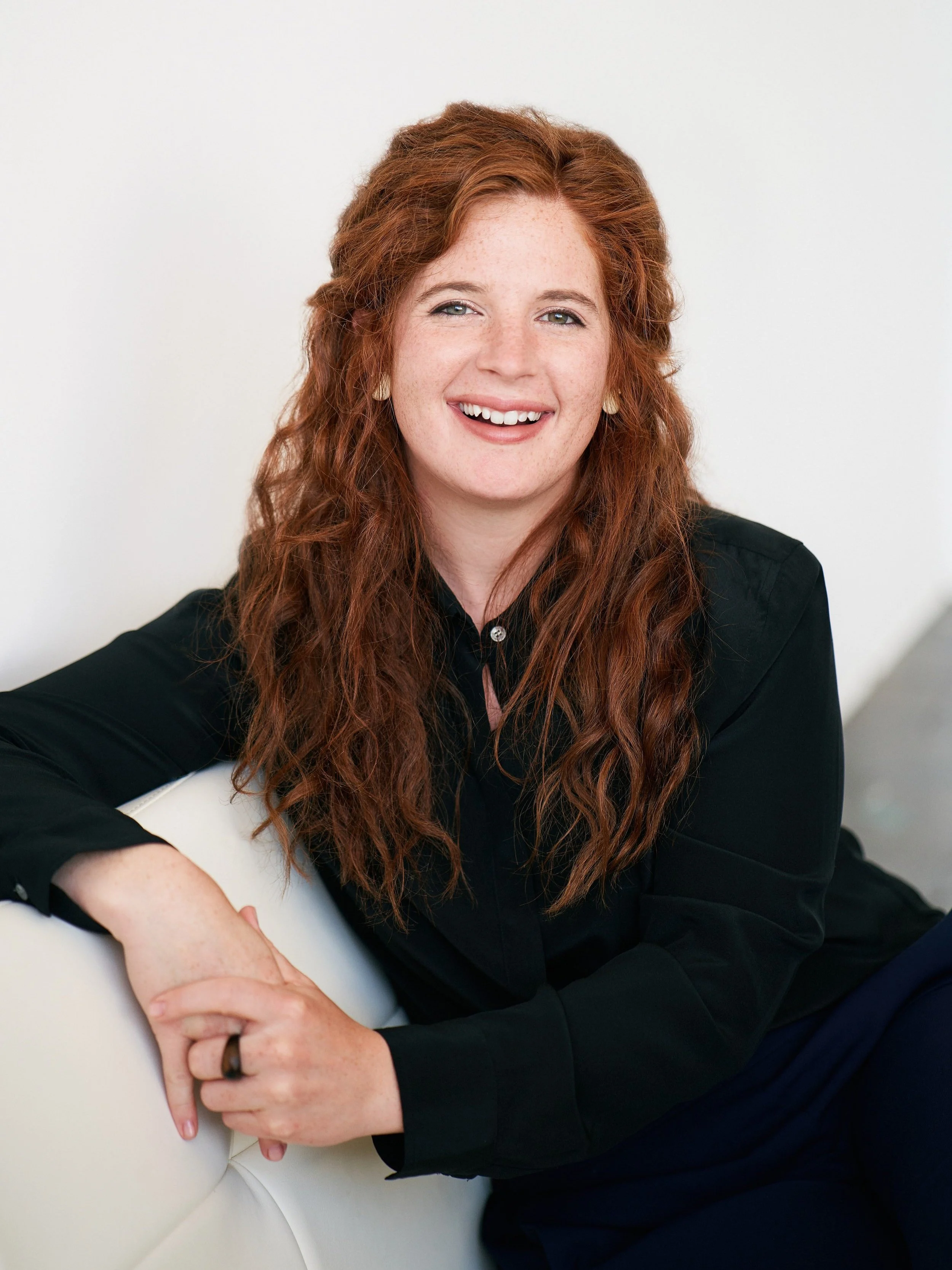 orporate portrait of a smiling executive seated on a white sofa in a bright office setting.