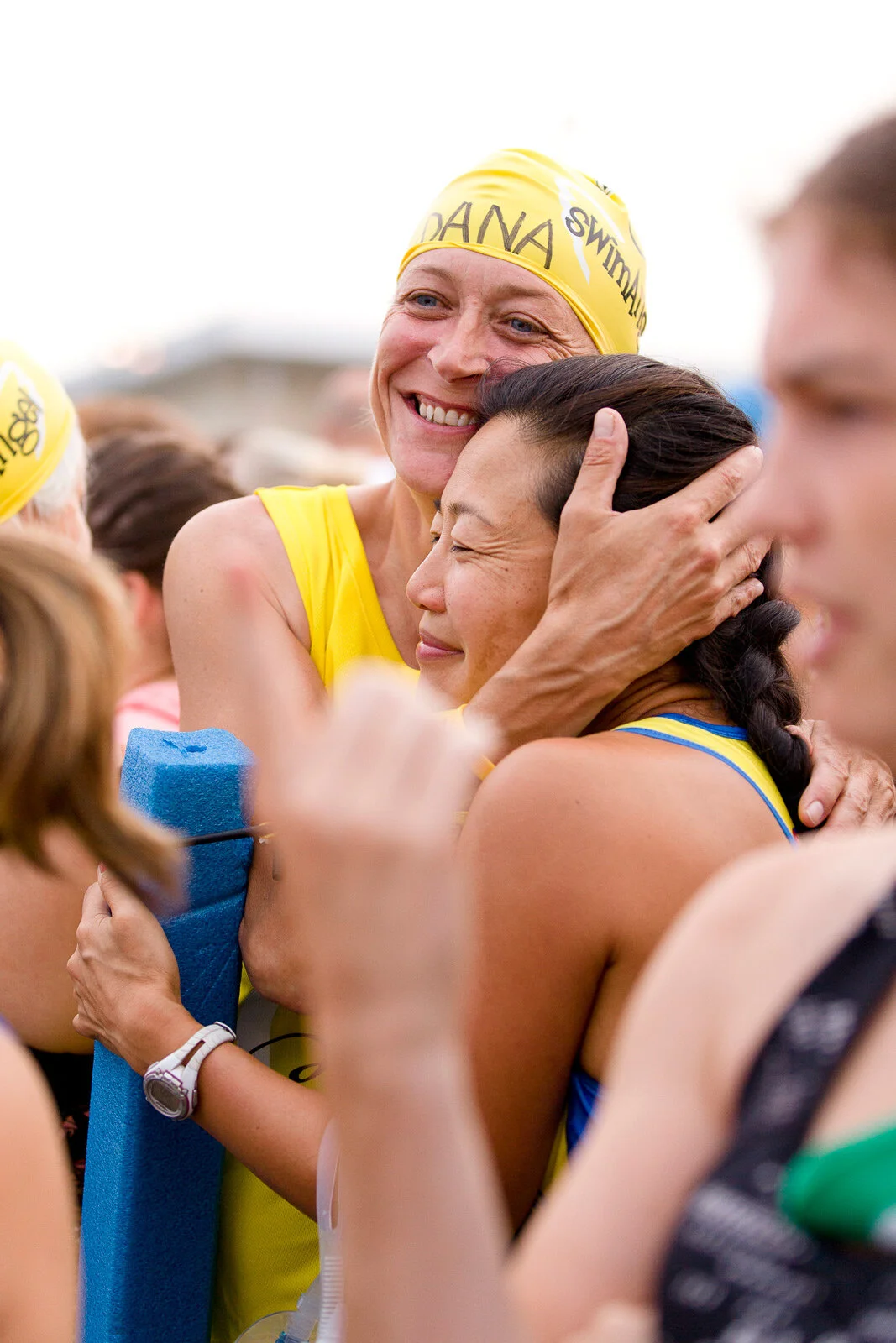 Two female athletes, wearing yellow and blue sport uniforms and swim caps, hug and celebrate at a sporting event, with one smiling warmly and the other closing her eyes.