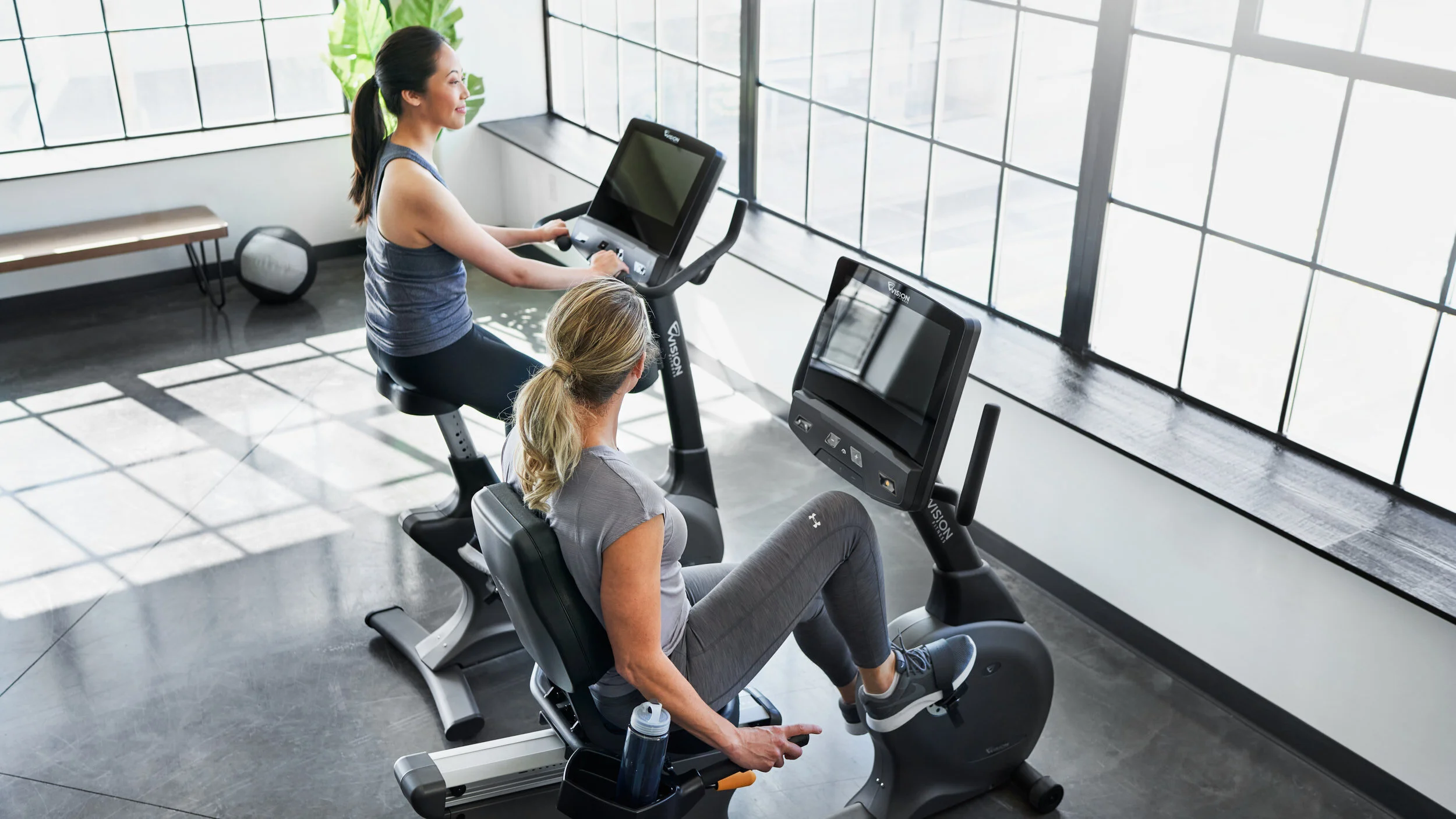 Two women exercising on stationary bikes in a brightly lit gym with large windows.