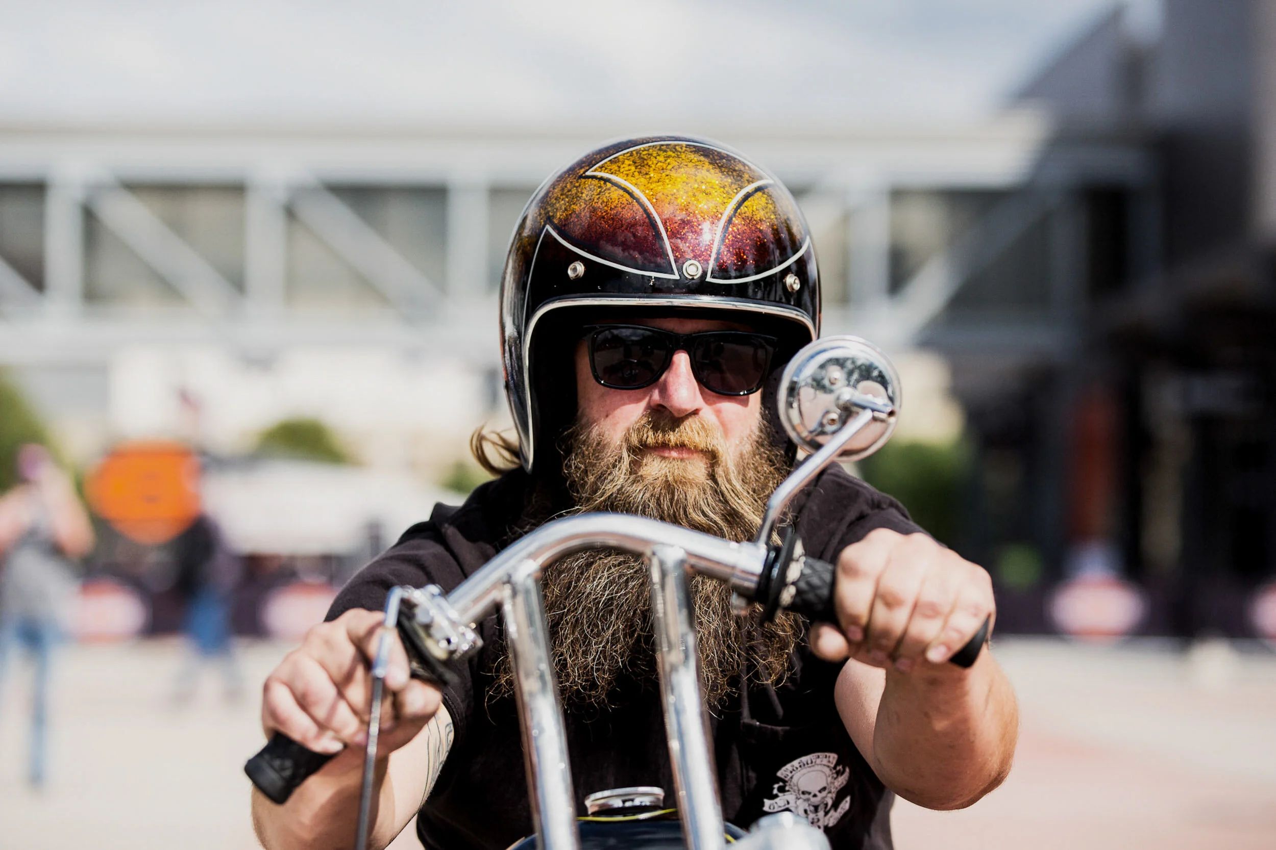 A man with a beard wearing a black helmet with a colorful design, sunglasses, and black clothing riding a motorcycle outdoors.