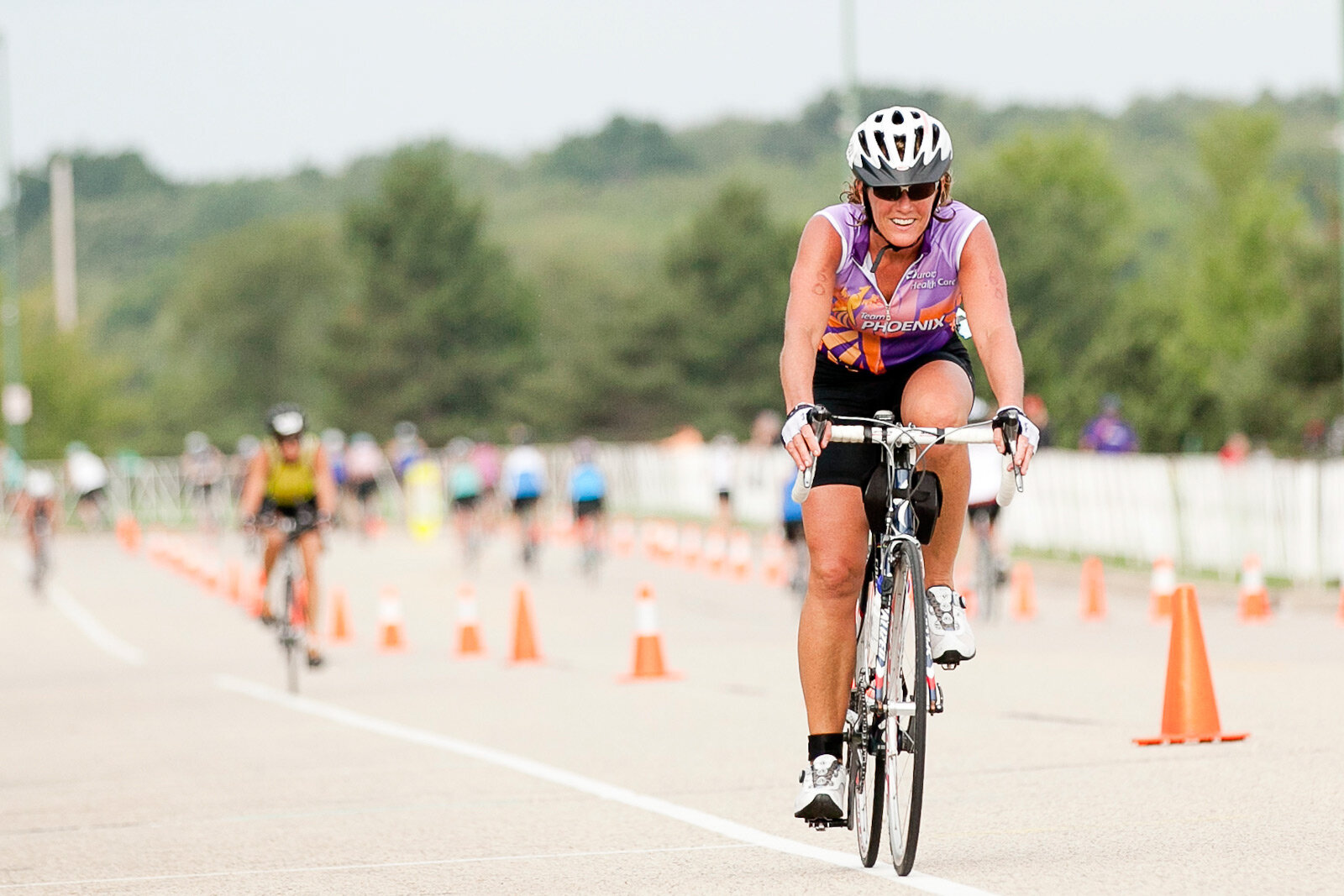 Female cyclist wearing helmet and sunglasses riding a bike during a race, with other cyclists and orange traffic cones in the background.