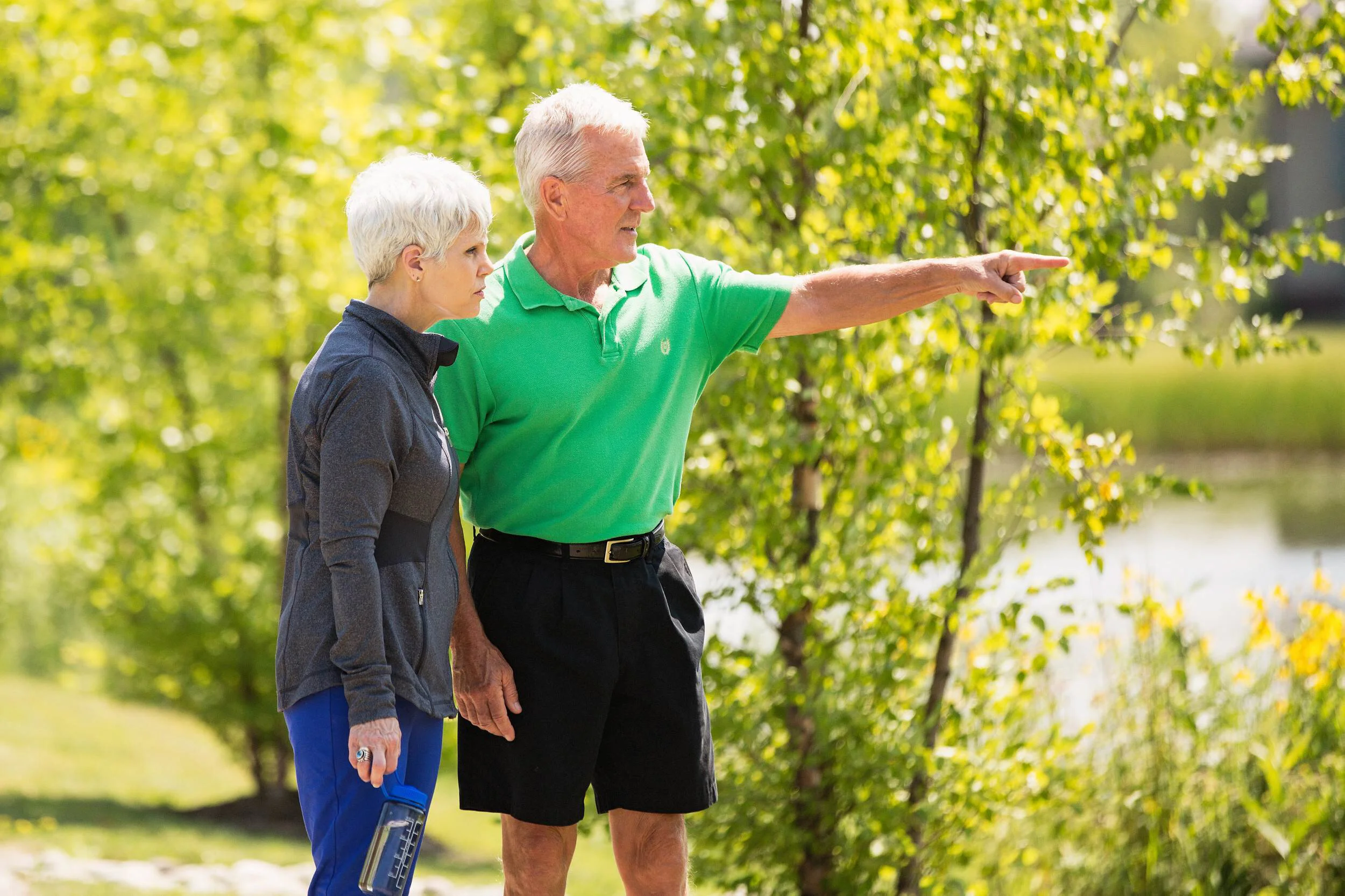 An elderly man in a green polo shirt and black shorts is pointing to the right while an elderly woman in a black jacket and blue pants looks on, holding a water bottle, outside in a park with green trees.