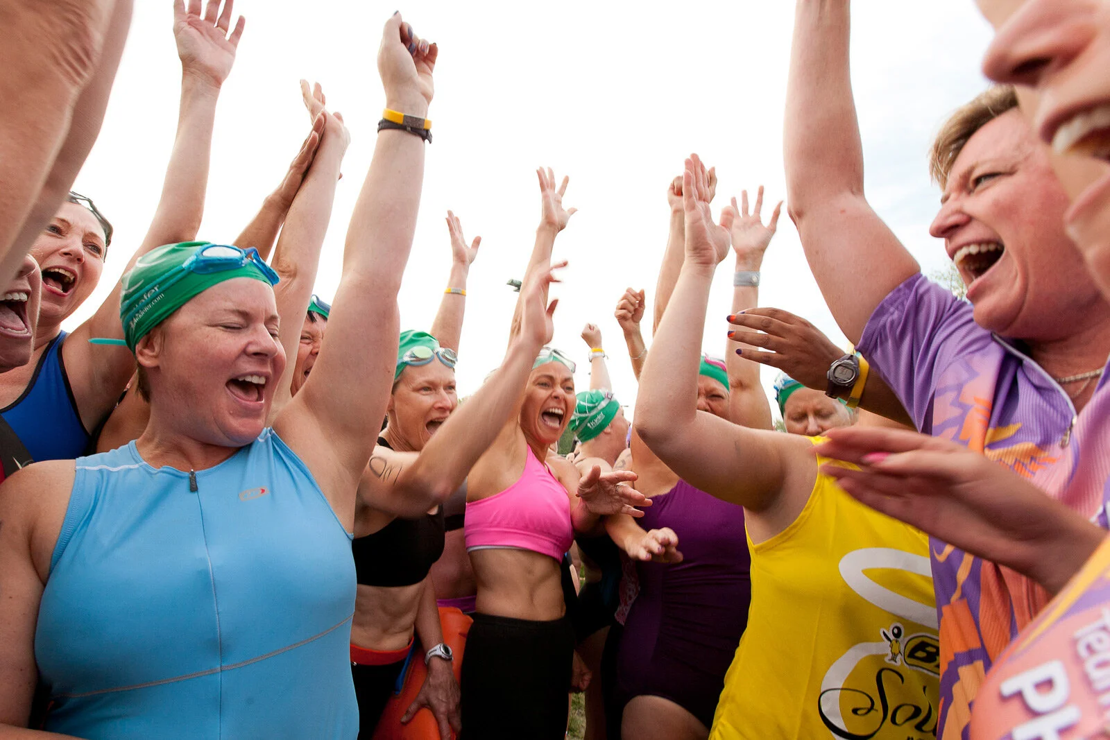 Group of female athletes wearing wetsuits and swim caps cheering and celebrating with hands raised at a sporting event.