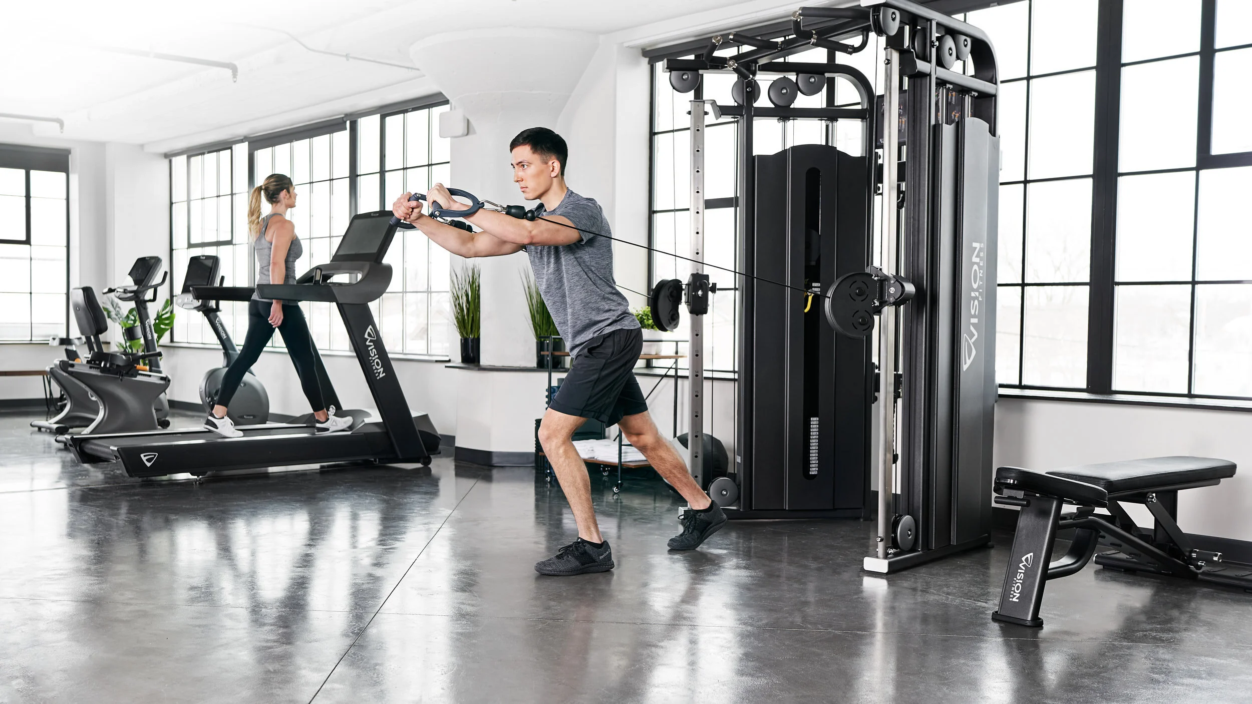 A man doing resistance band exercises in a bright gym with large windows, while a woman walks on a treadmill in the background.