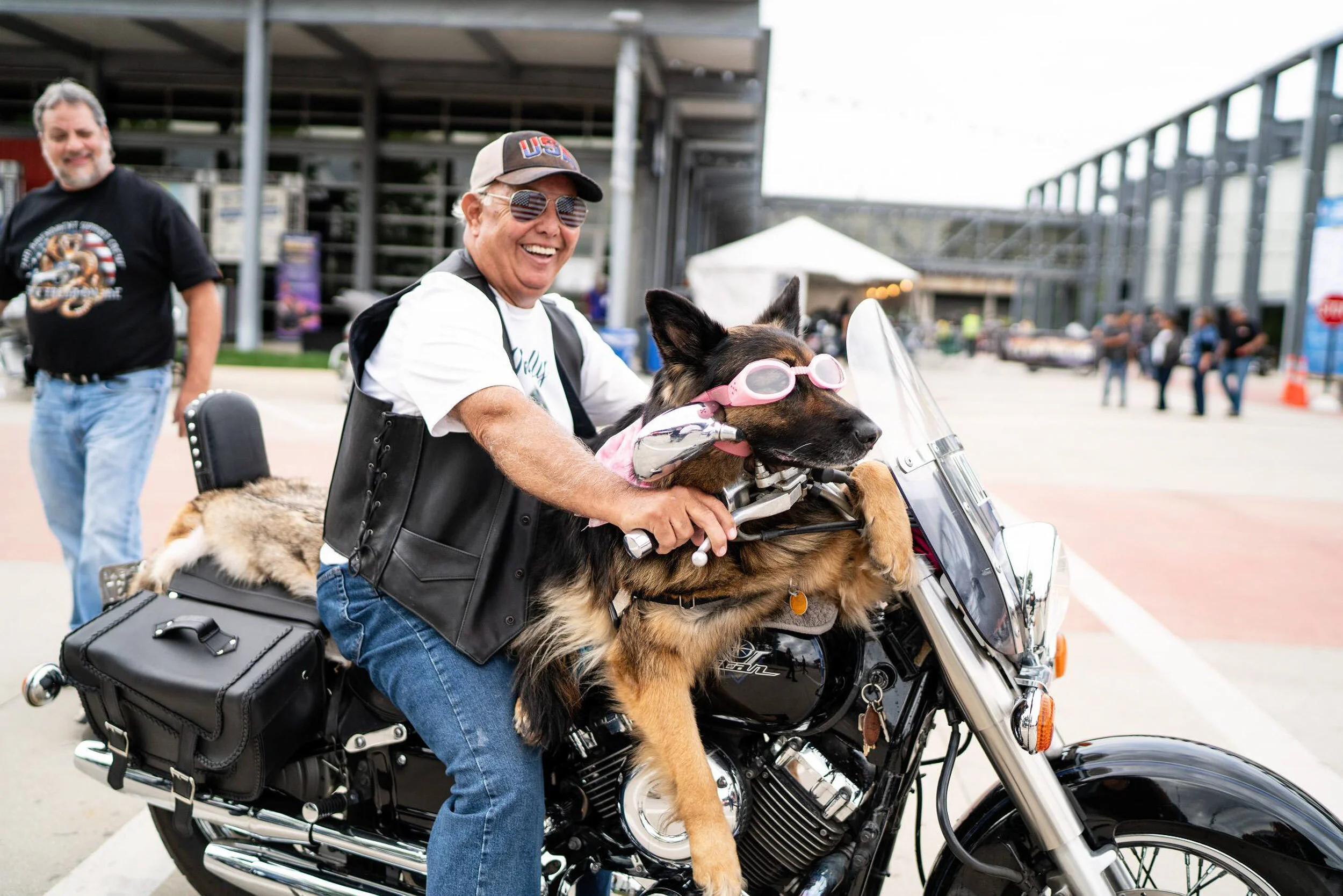 A man with a beard riding a motorcycle with a dog in front wearing pink goggles, and a man in the background smiling at an outdoor event.