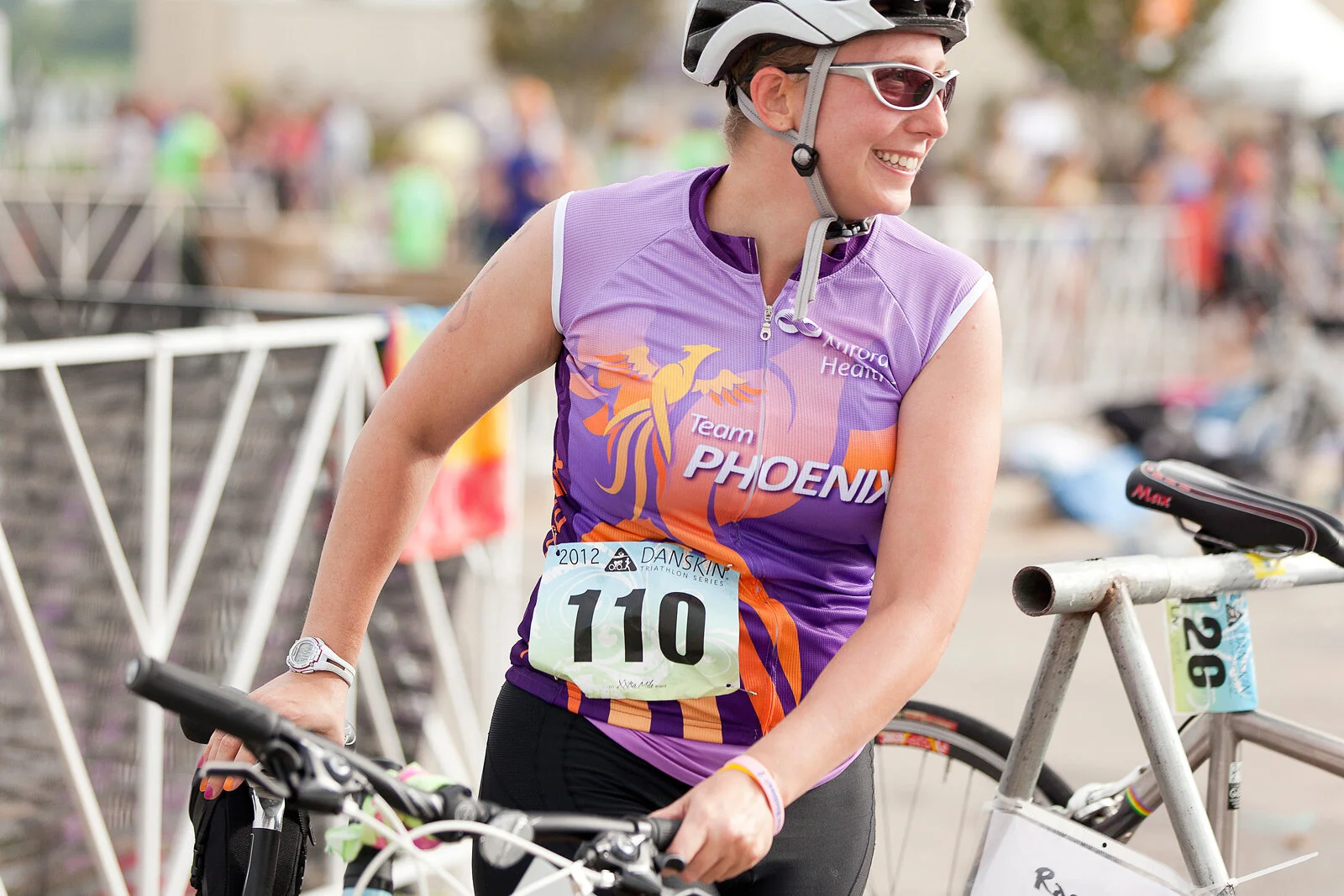 A woman wearing a purple and orange triathlon jersey, a helmet, and sunglasses, smiling while holding a bicycle at a race event with a race number 110.