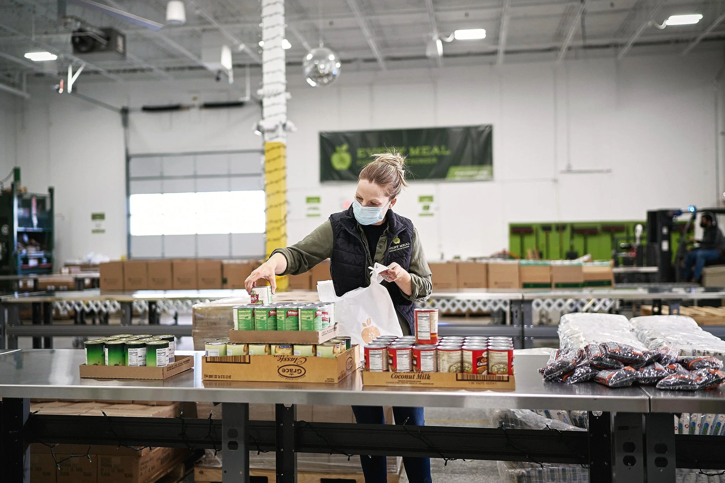 Masked volunteer organizing canned goods on a packing table at Every Meal Minnesota warehouse.