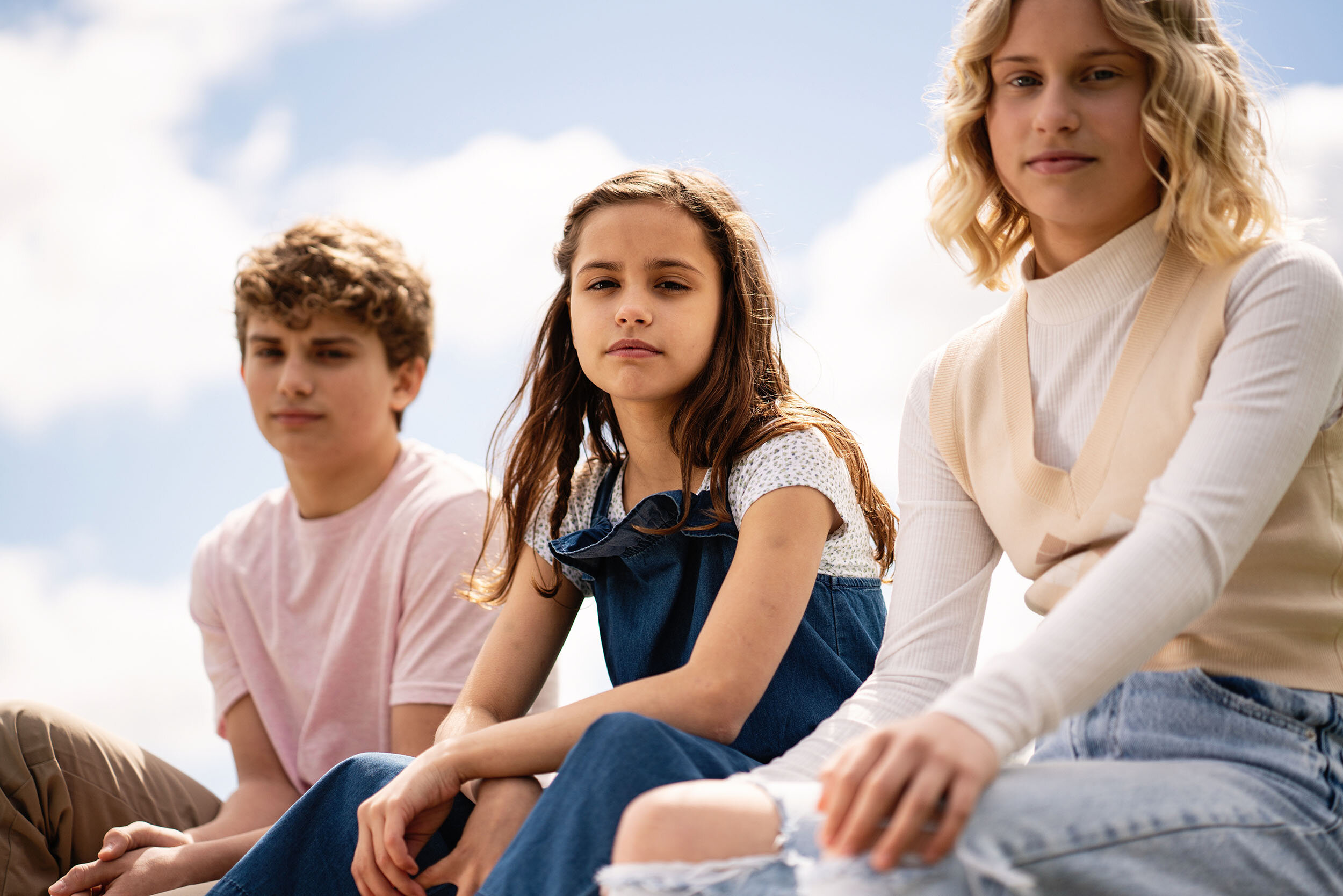 Three teenagers sitting outdoors on a sunny day with a partly cloudy sky in the background.