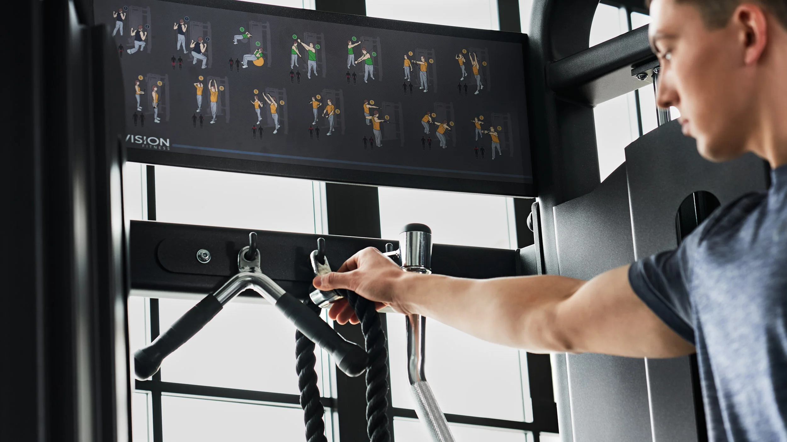 A person adjusting a piece of gym equipment at a fitness center, with a screen displaying illustrated workout instructions in the background.
