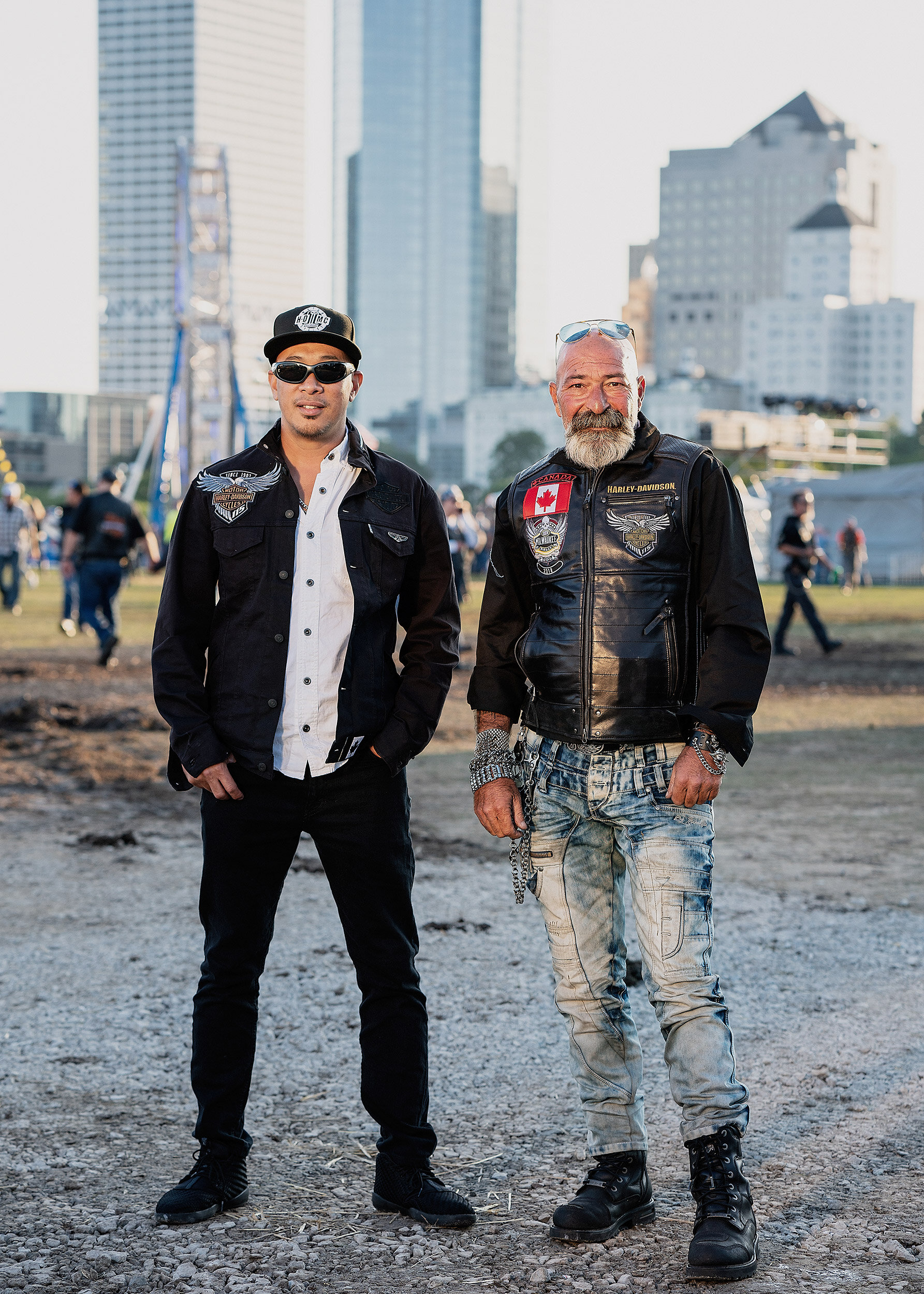 Two men standing outdoors in front of a cityscape, one wearing sunglasses, a white shirt, and a black jacket, the other with a beard, sunglasses on his head, and a leather vest, both dressed in casual clothing and biker attire.