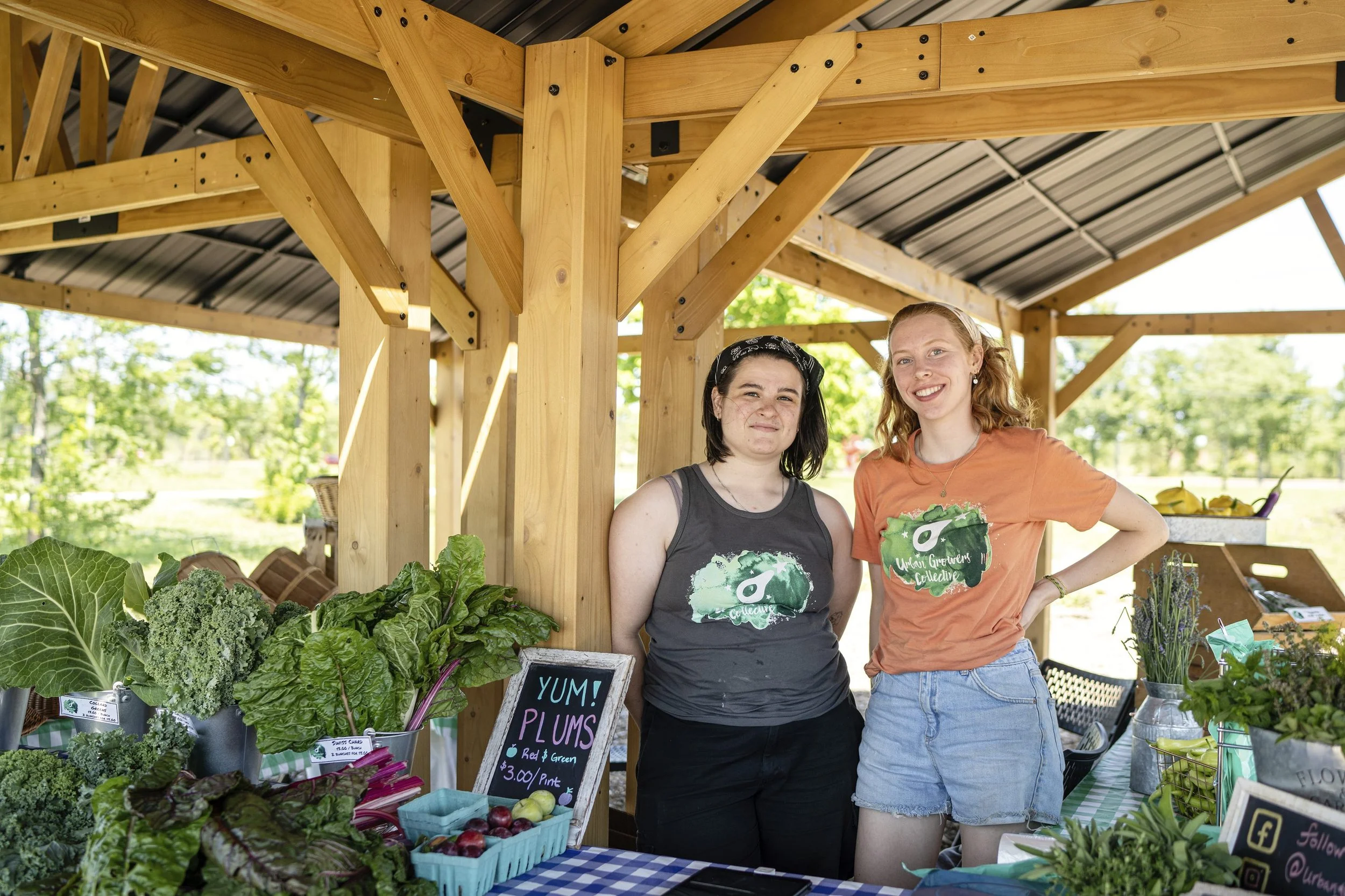 Two volunteers standing behind a farm stand displaying fresh greens and produce at a community farmers market.