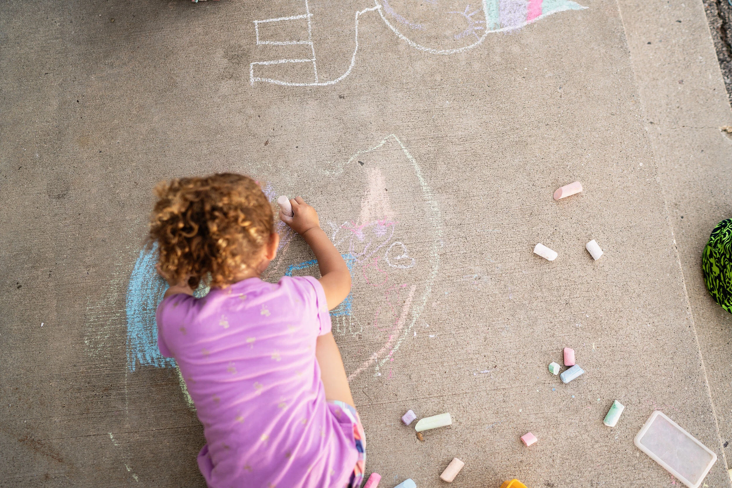 A young girl with curly hair drawing with chalk on a concrete sidewalk, creating colorful chalk art and messages.