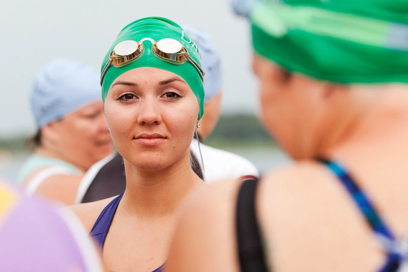Close-up of a young woman in a green swim cap with goggles on her forehead, surrounded by other swimmers wearing swim caps, outdoors during daytime.