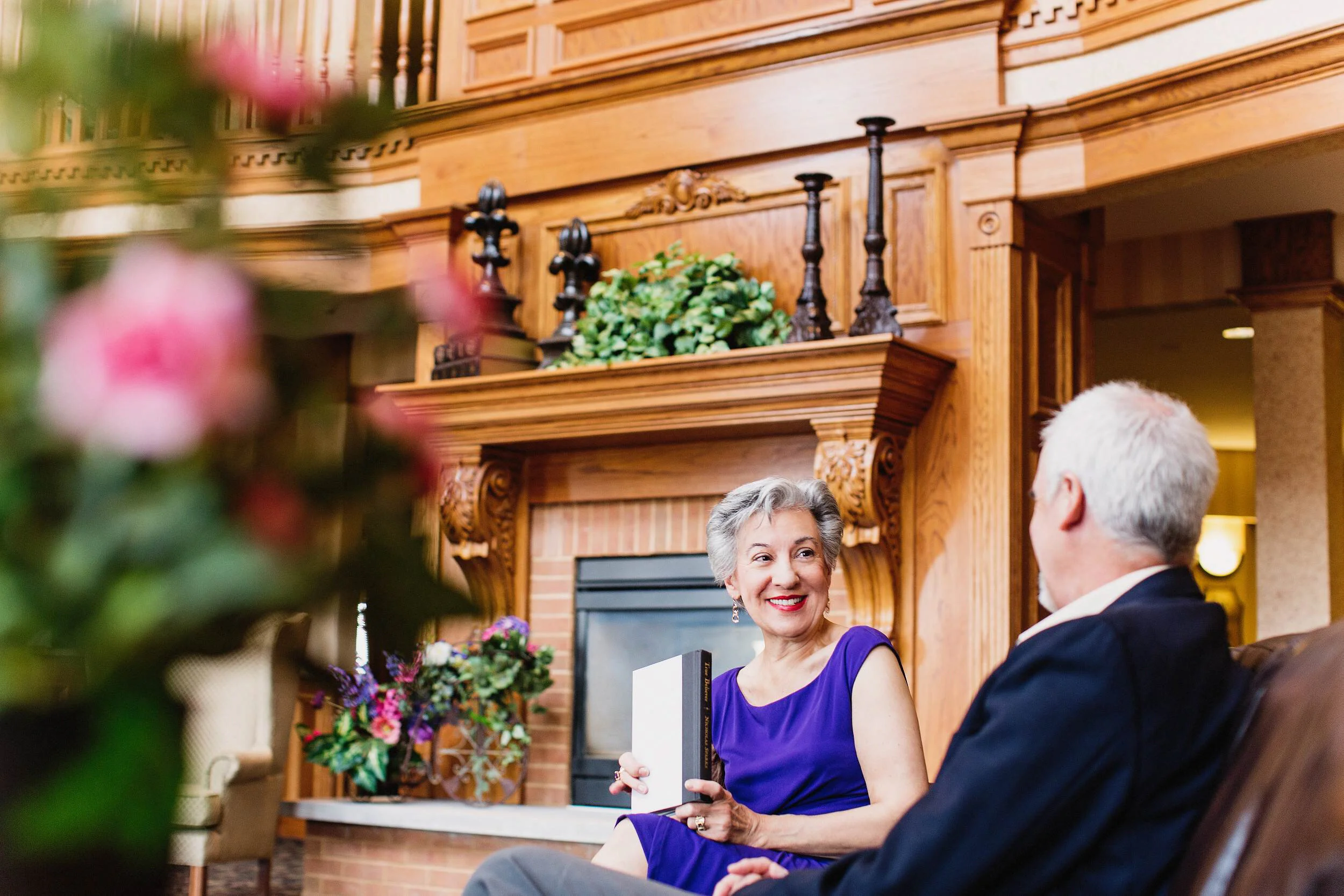 A woman in a purple dress smiling and holding a book, talking to a man with white hair, in a warmly lit room with a brick fireplace and wood paneling.
