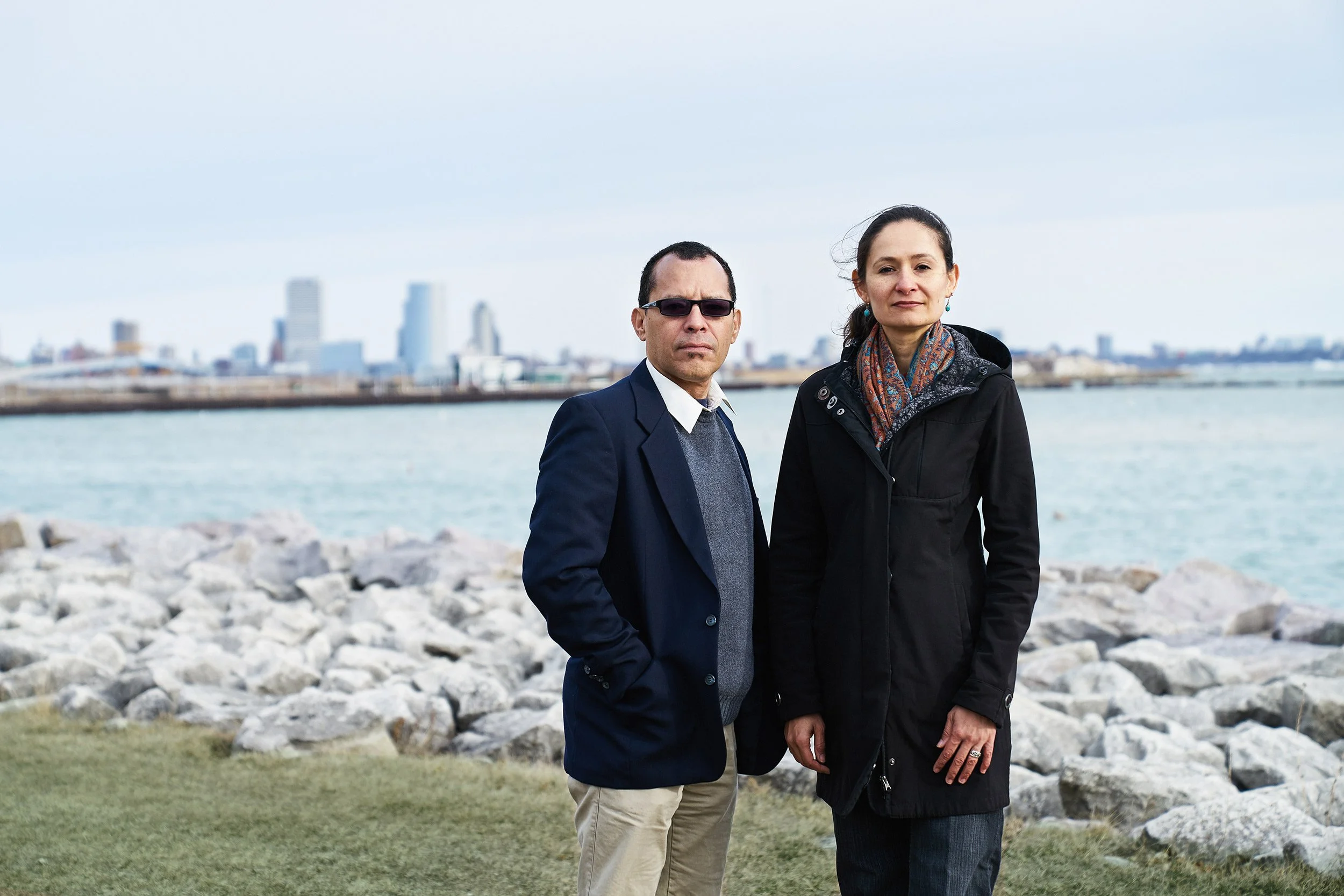 Two community leaders standing together along a rocky lakefront with a city skyline in the background.