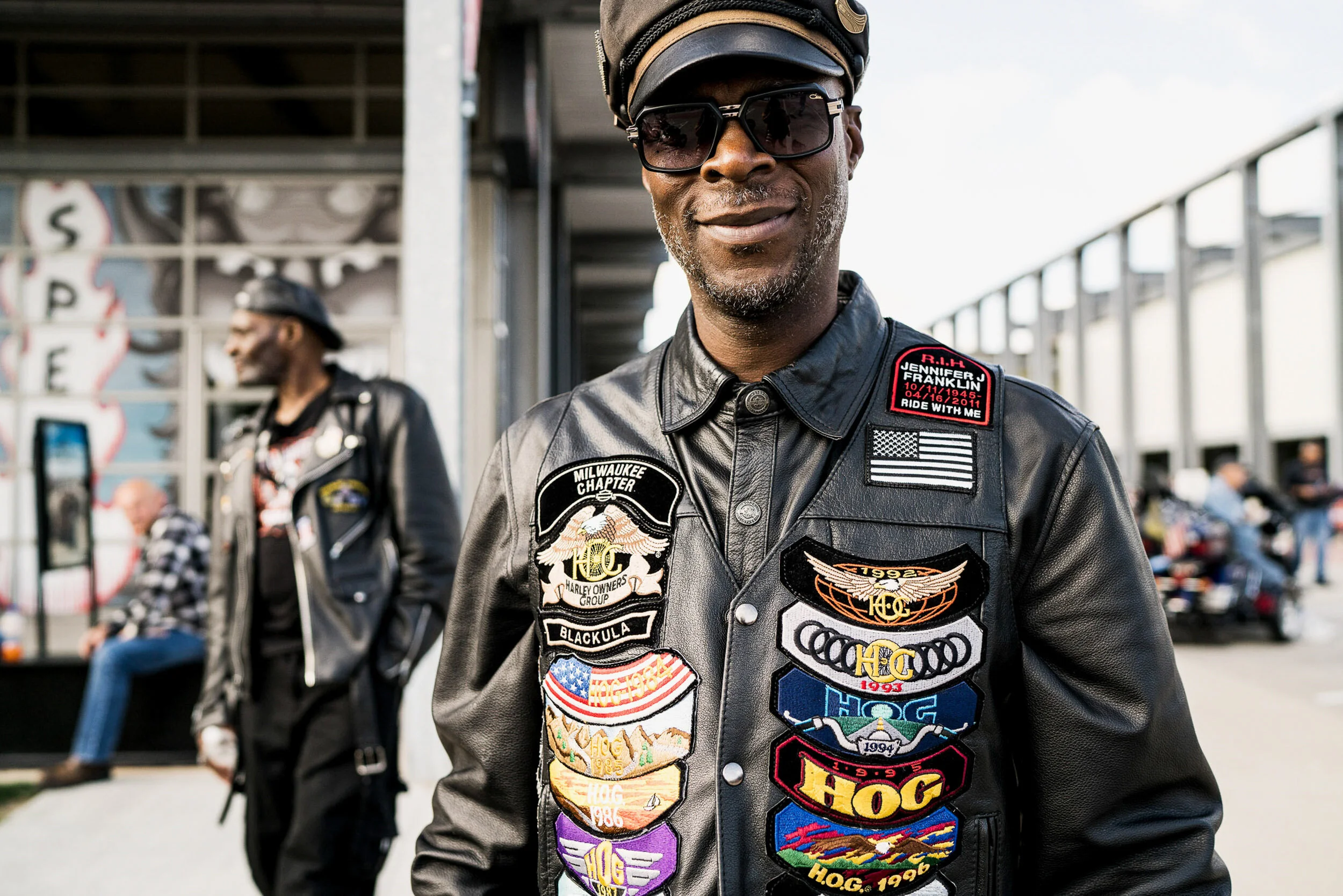 A smiling man wearing sunglasses and a black leather jacket decorated with apocalyptic patches, standing outdoors with people and motorcycles in the background.