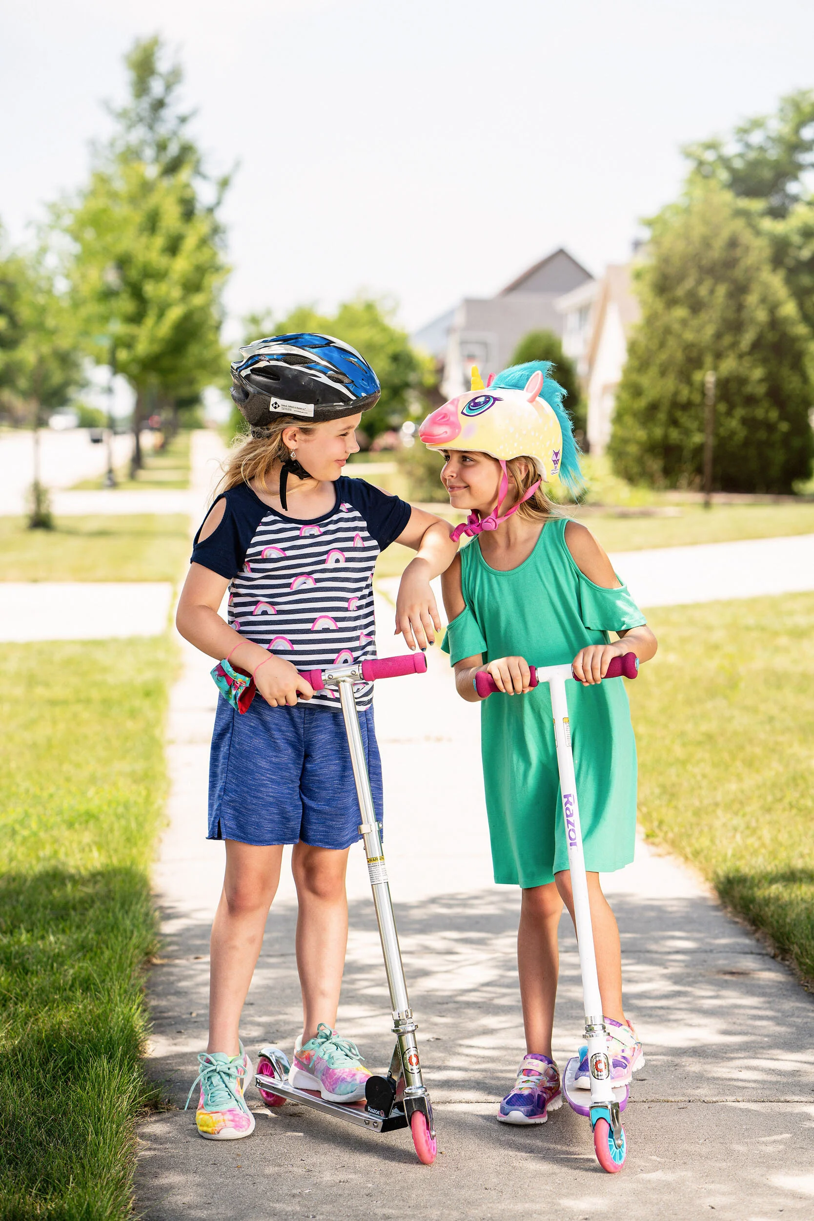 Two young girls standing on a sidewalk, wearing helmets, and holding scooters, talking and smiling at each other in a residential neighborhood.