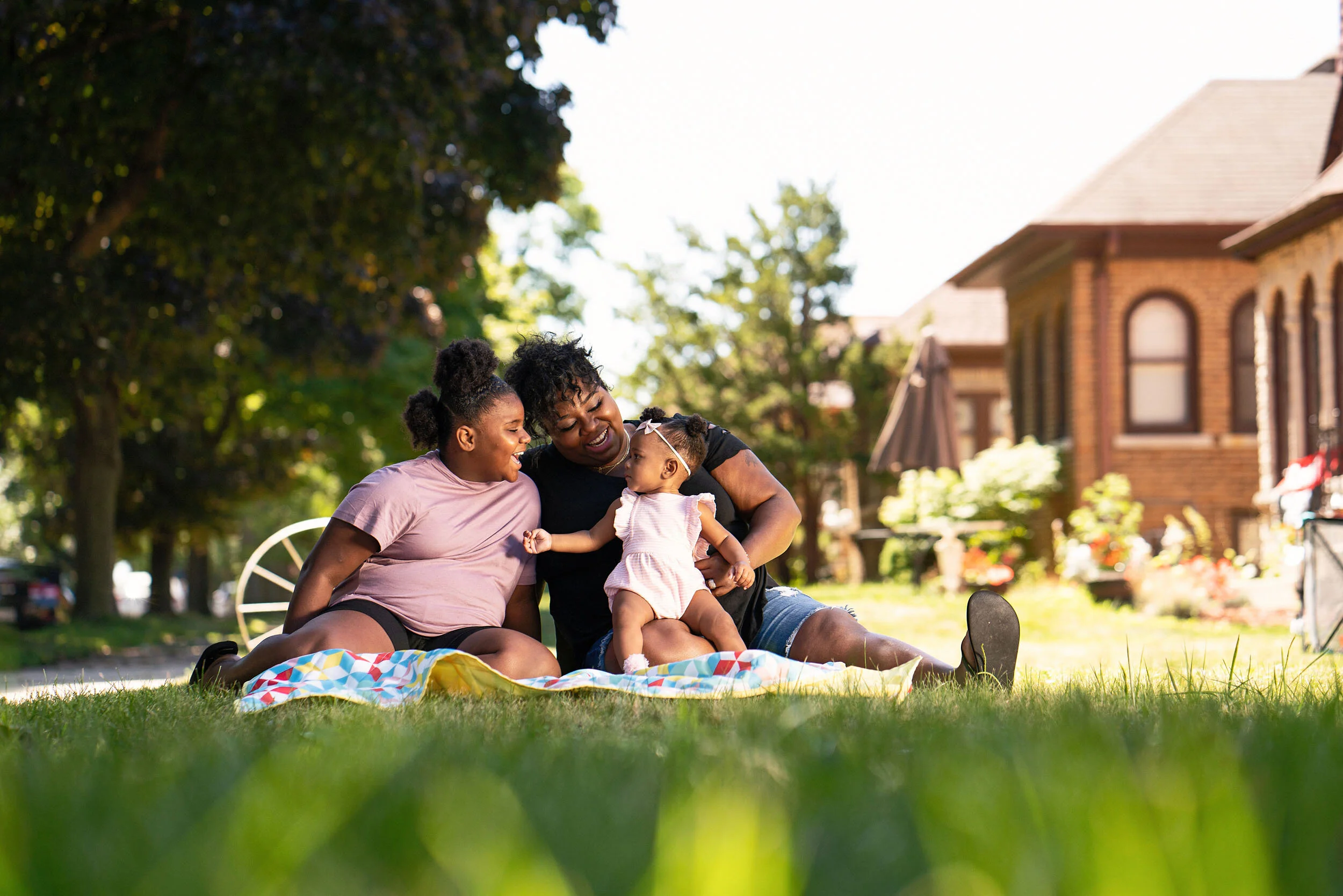 A woman and two young girls sitting on a blanket on the grass in a residential neighborhood, smiling and playing with a baby girl, surrounded by trees and houses.