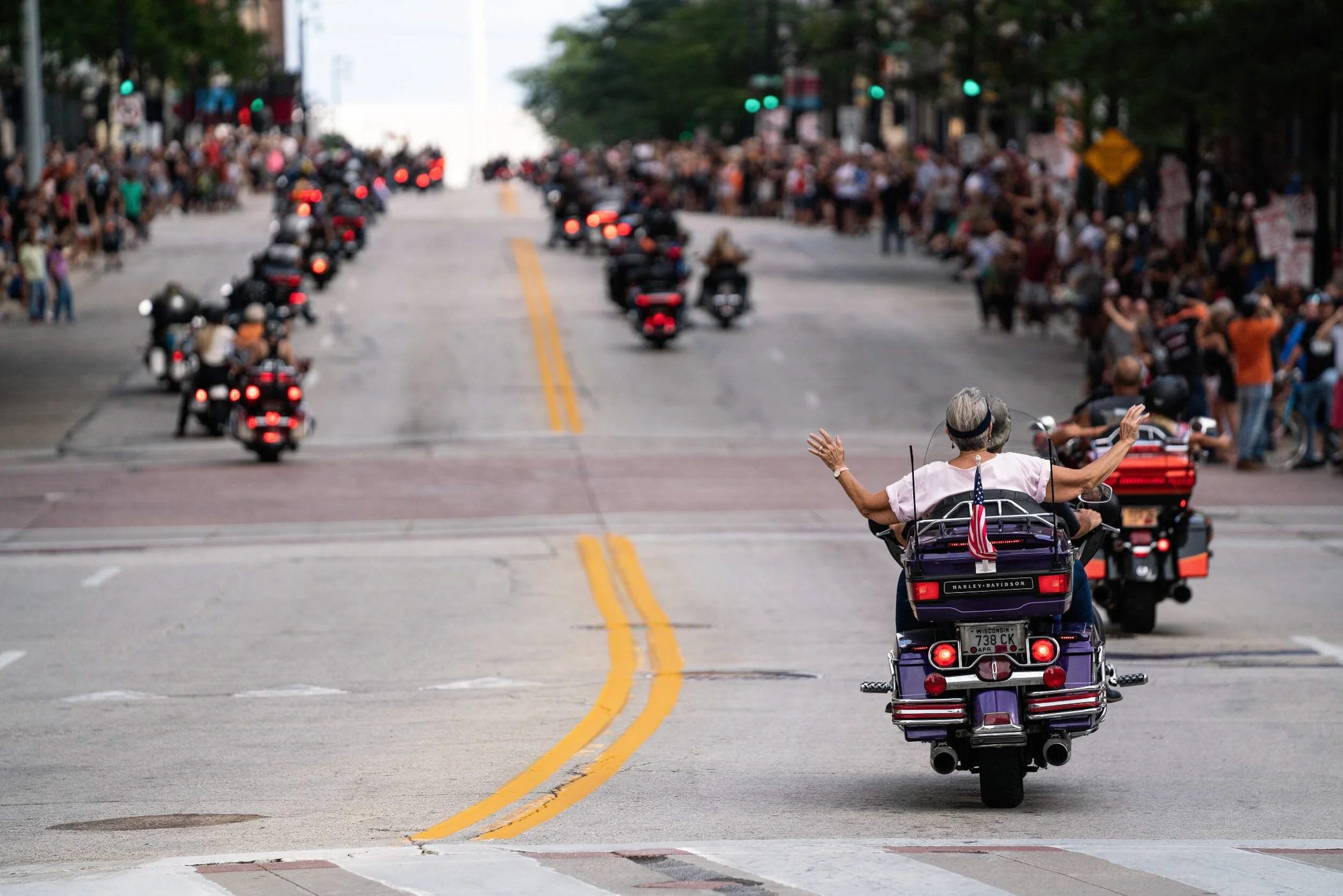 A woman riding a purple Harley-Davidson motorcycle with her arms raised, leading a procession of motorcycles and people on the street during a parade or rally, with spectators lining the sidewalk.