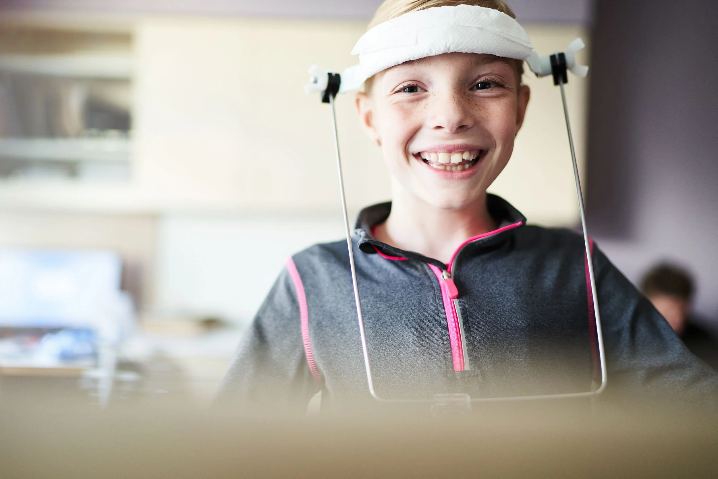 A smiling young girl with a gap-tooth smile, wearing a grey zip-up jacket with pink accents, sitting at a desk with a clear face shield on her head.
