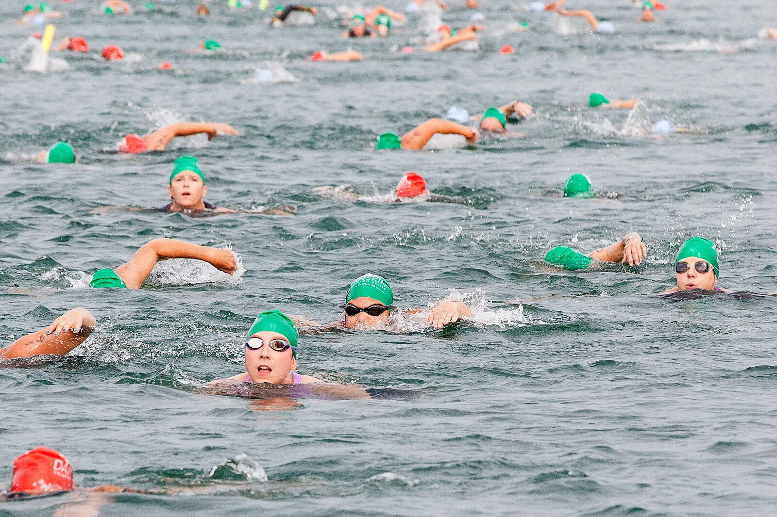 Swimmers participating in a competitive open water swimming race, wearing green and red swim caps and goggles, in a large body of water.