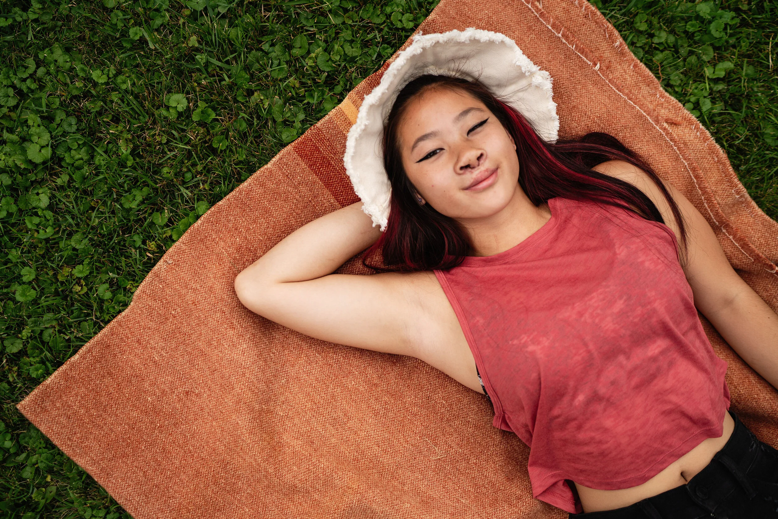 A young girl with long dark hair and red highlights lies on a terracotta-colored mat on green grass, wearing a red sleeveless top and a white wide-brimmed hat with a frayed edge, smiling at the camera.