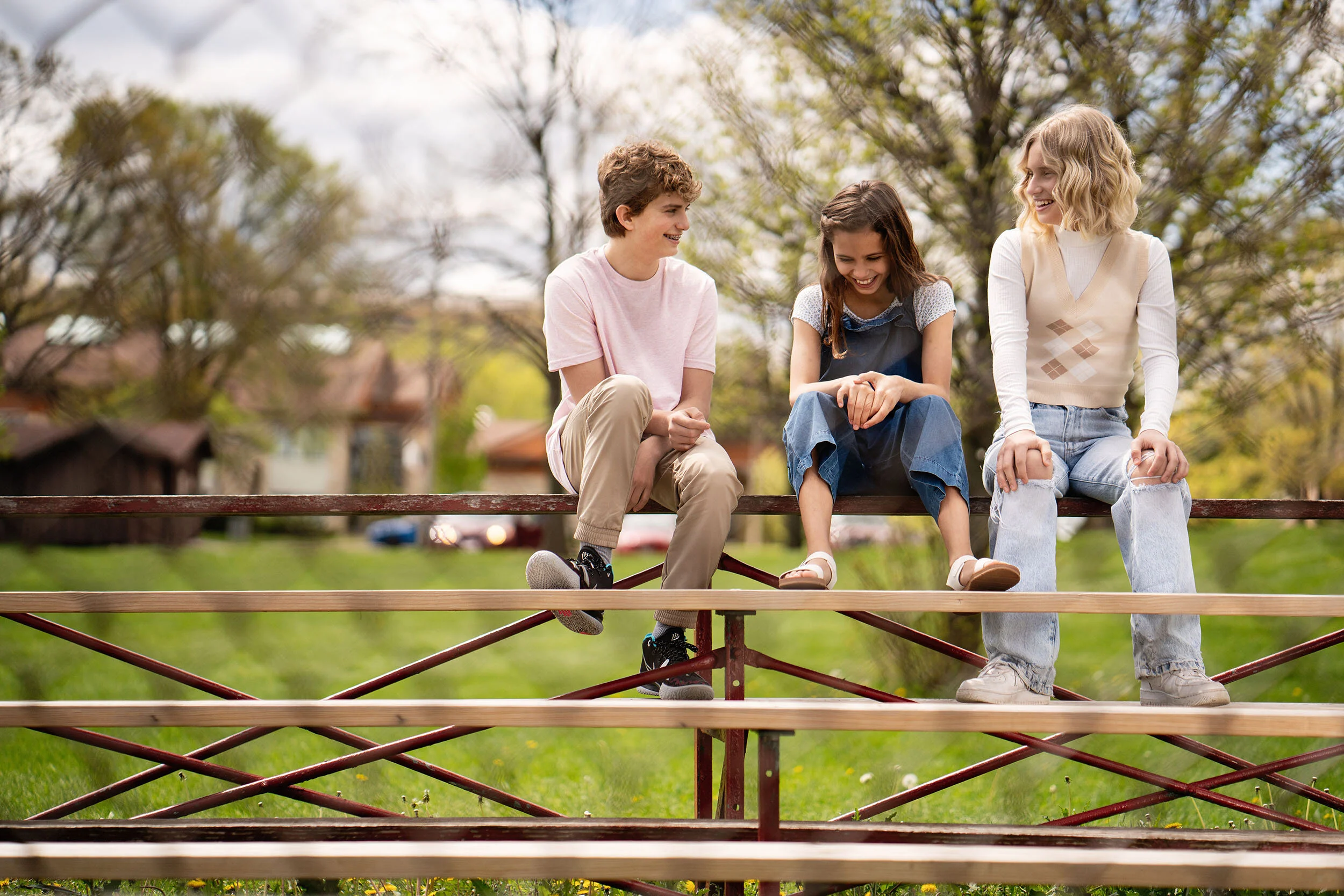 Three children sitting on a metal and wood bench outdoors, smiling and talking, with trees and houses in the background.