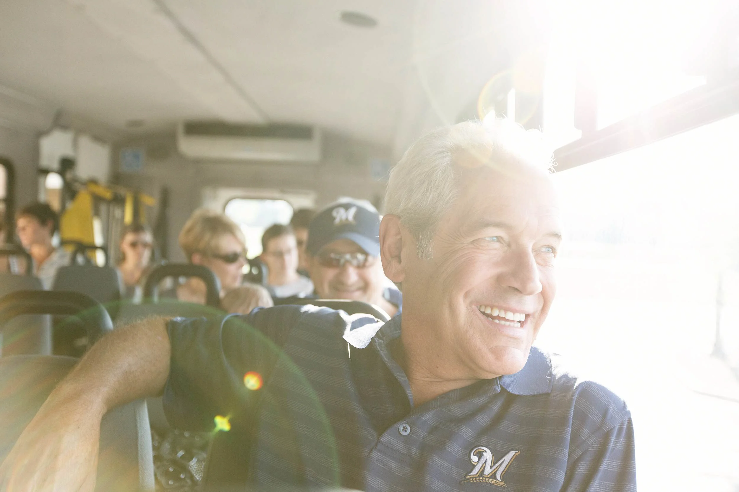 A smiling older man with gray hair, wearing a navy blue polo shirt with an embroidered 'M' on it, sitting on a bus with other passengers, enjoying sunlight coming through the window.