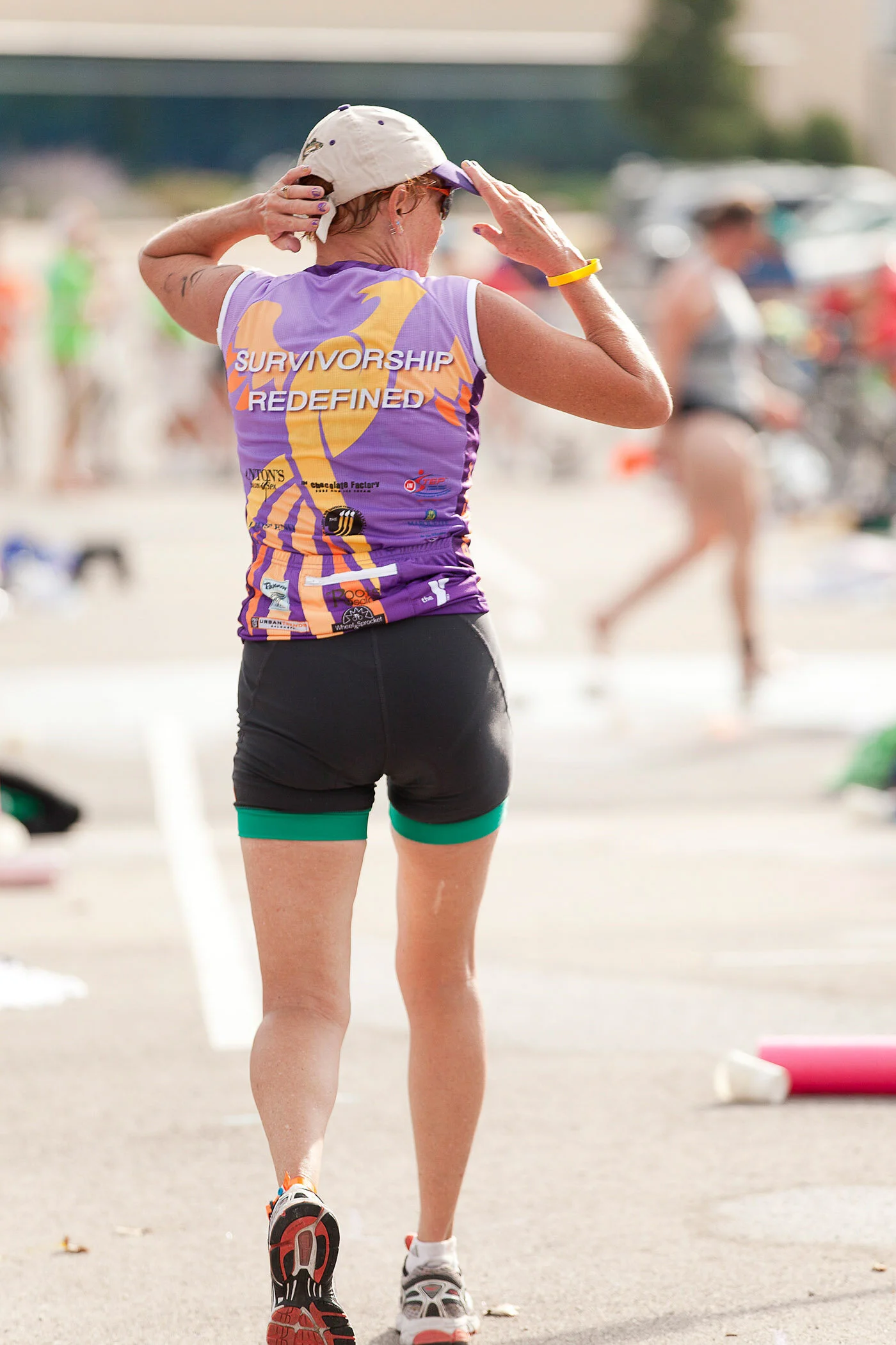 A woman in athletic clothing, wearing a purple tank top and black shorts, stands on a street during a race or event, adjusting her cap.
