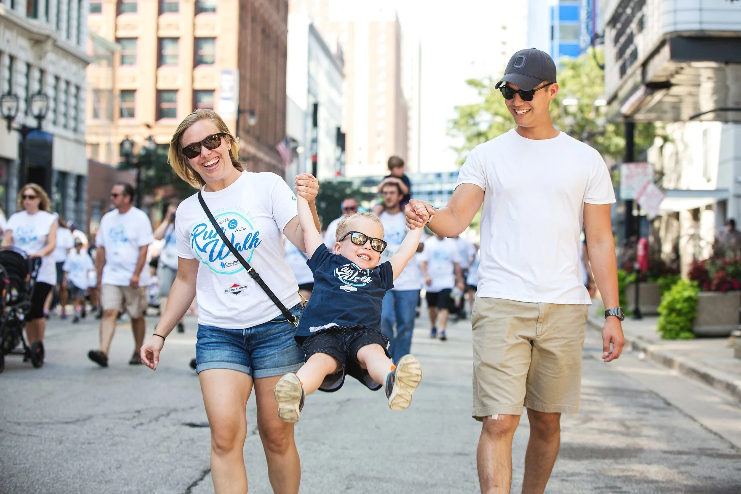 Family participating in a downtown charity run and walk fundraiser, smiling while holding their child between them.