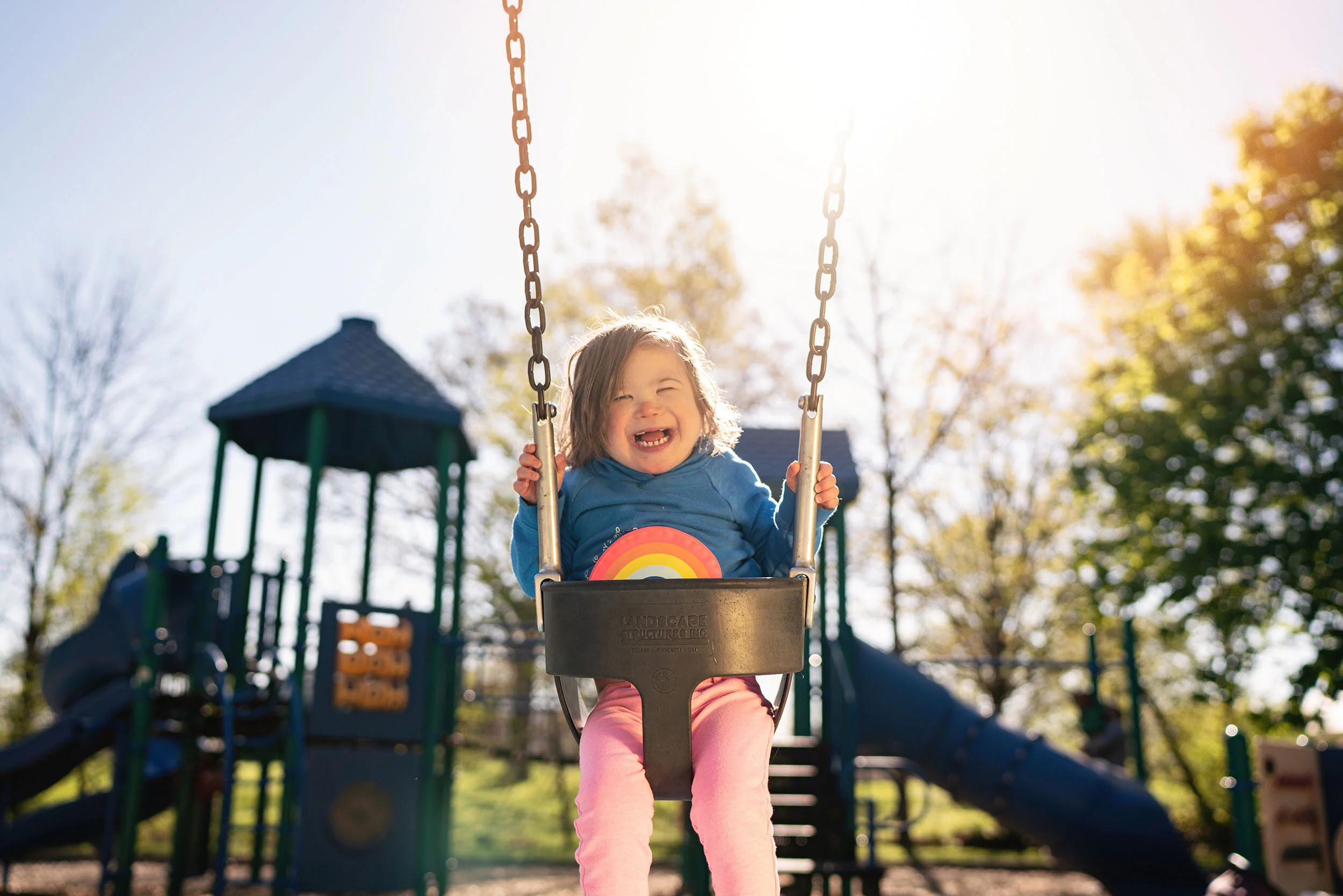 A young girl is happily swinging on a playground swing set, with sunlight shining behind her and trees in the background.