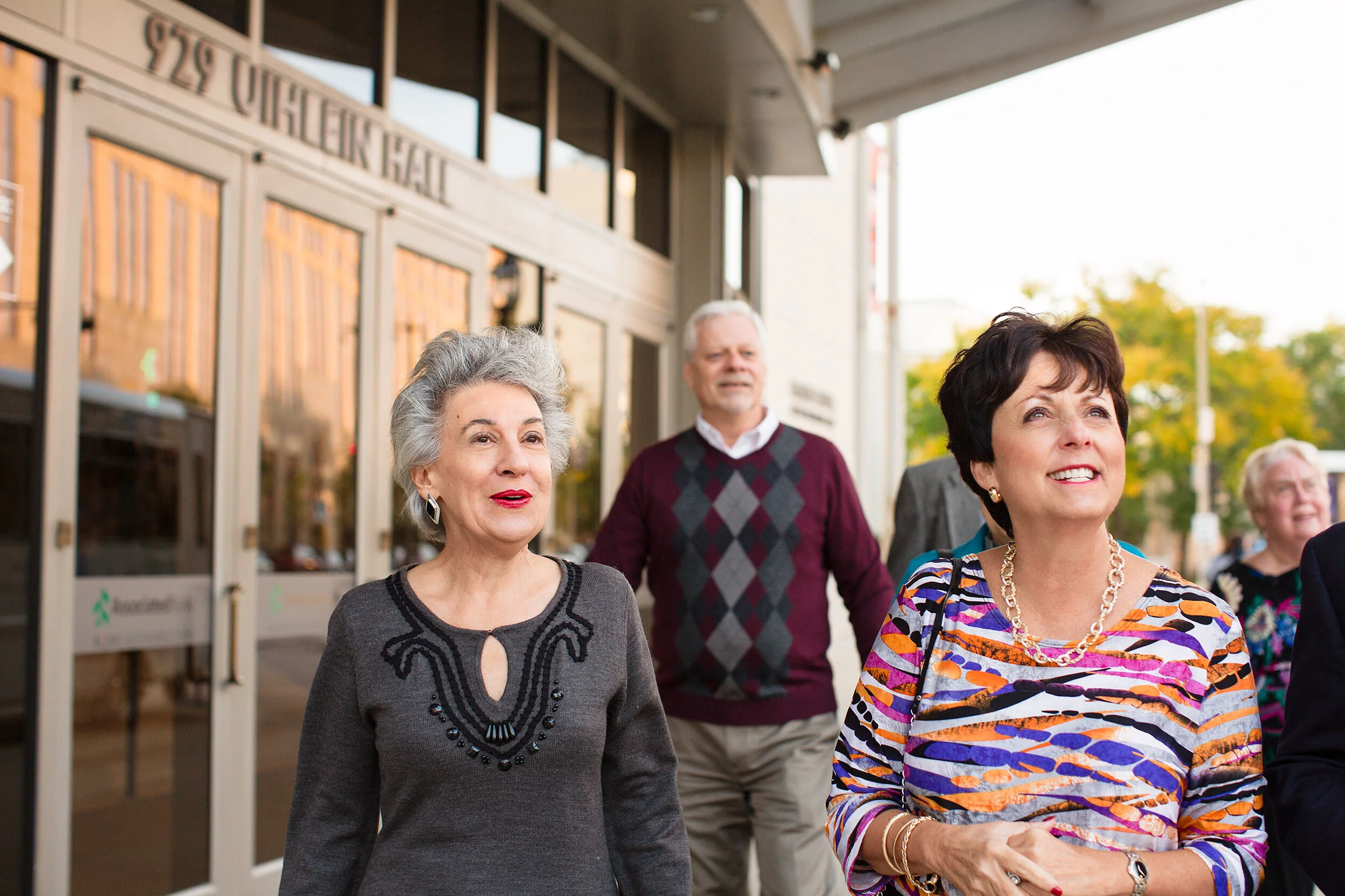 Group of older adults walking and smiling outside a building with glass doors.