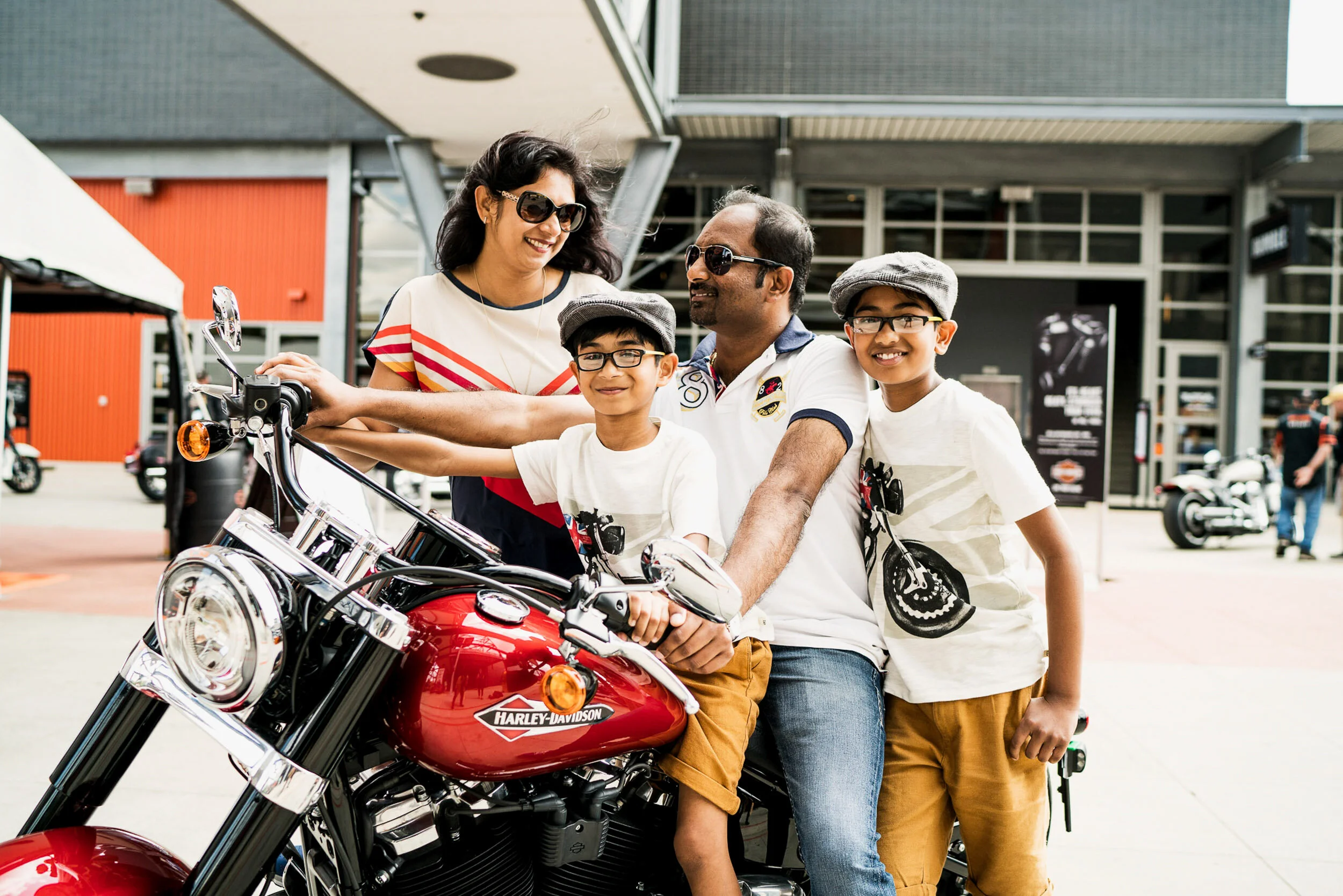 A family of four, including a mother, father, and two young boys, is gathered around a red Harley-Davidson motorcycle outside a modern building. They are smiling and appear excited and happy, with the father sitting on the motorcycle and the boys sta
