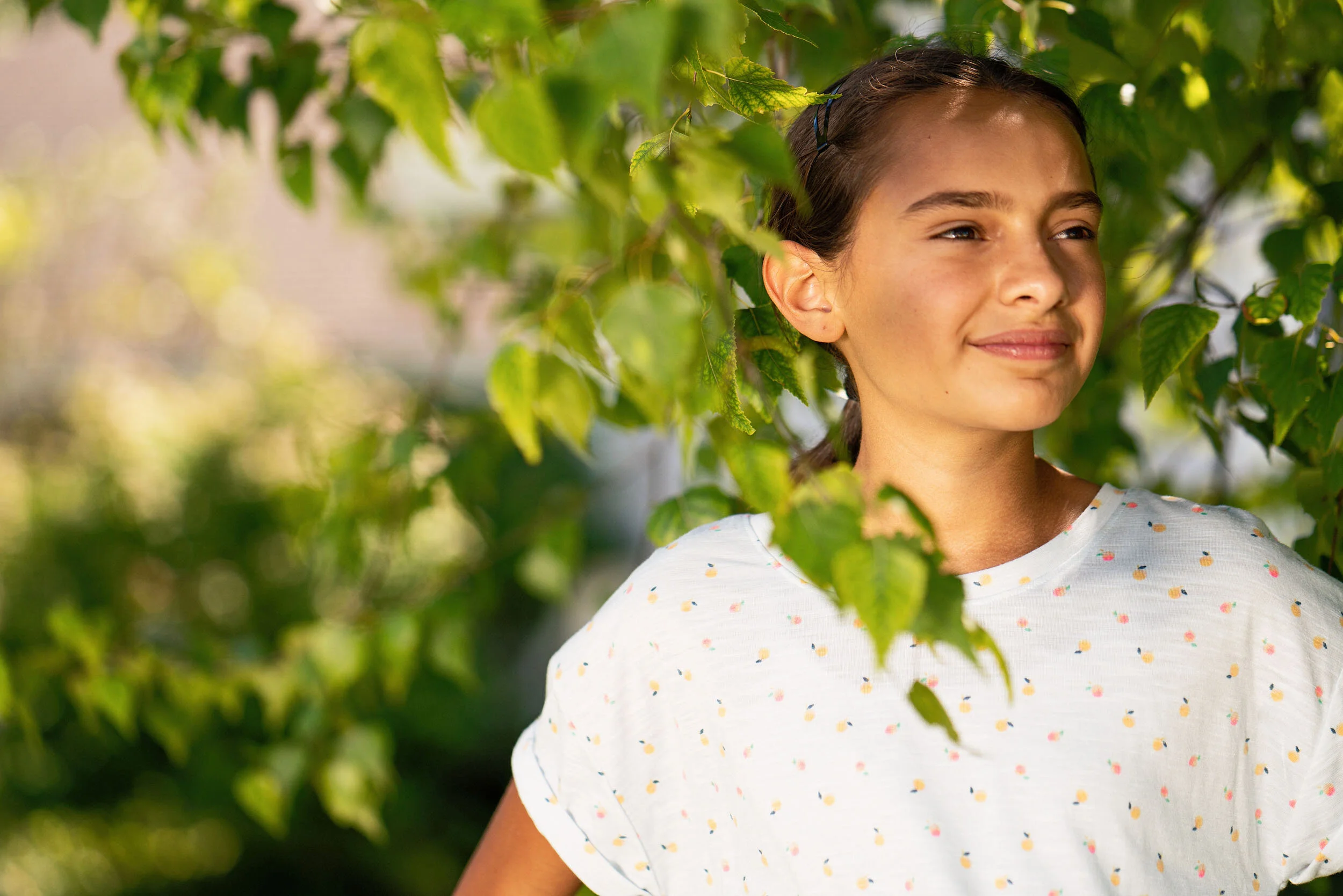 A young woman with brown hair and a white t-shirt with colorful dots, standing outdoors among green leaves, smiling softly and looking into the distance.