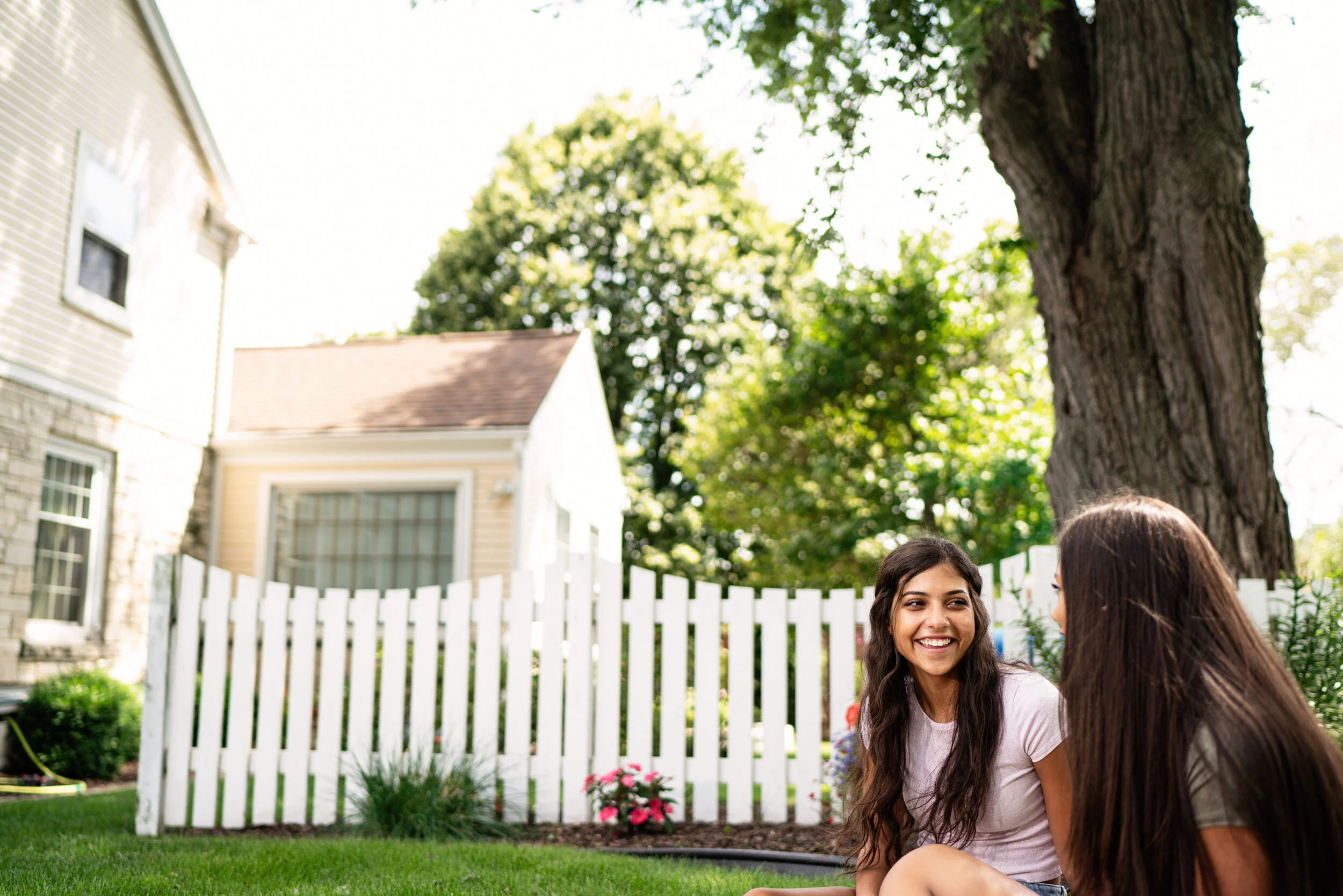 Two young women sitting on grass in a backyard, smiling and talking, with a large tree, white fence, and houses in the background.