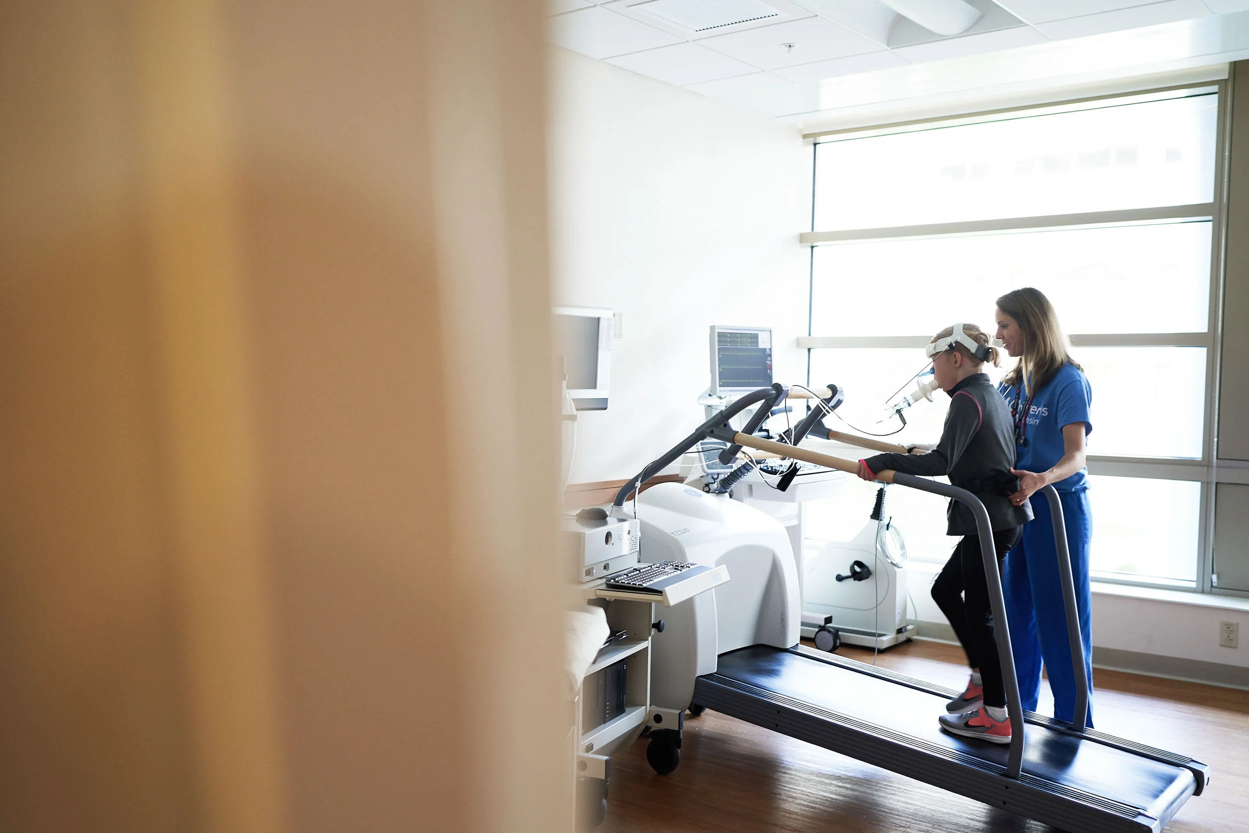 A nurse assisting a young patient walking on a treadmill in a hospital with medical monitors in the background.