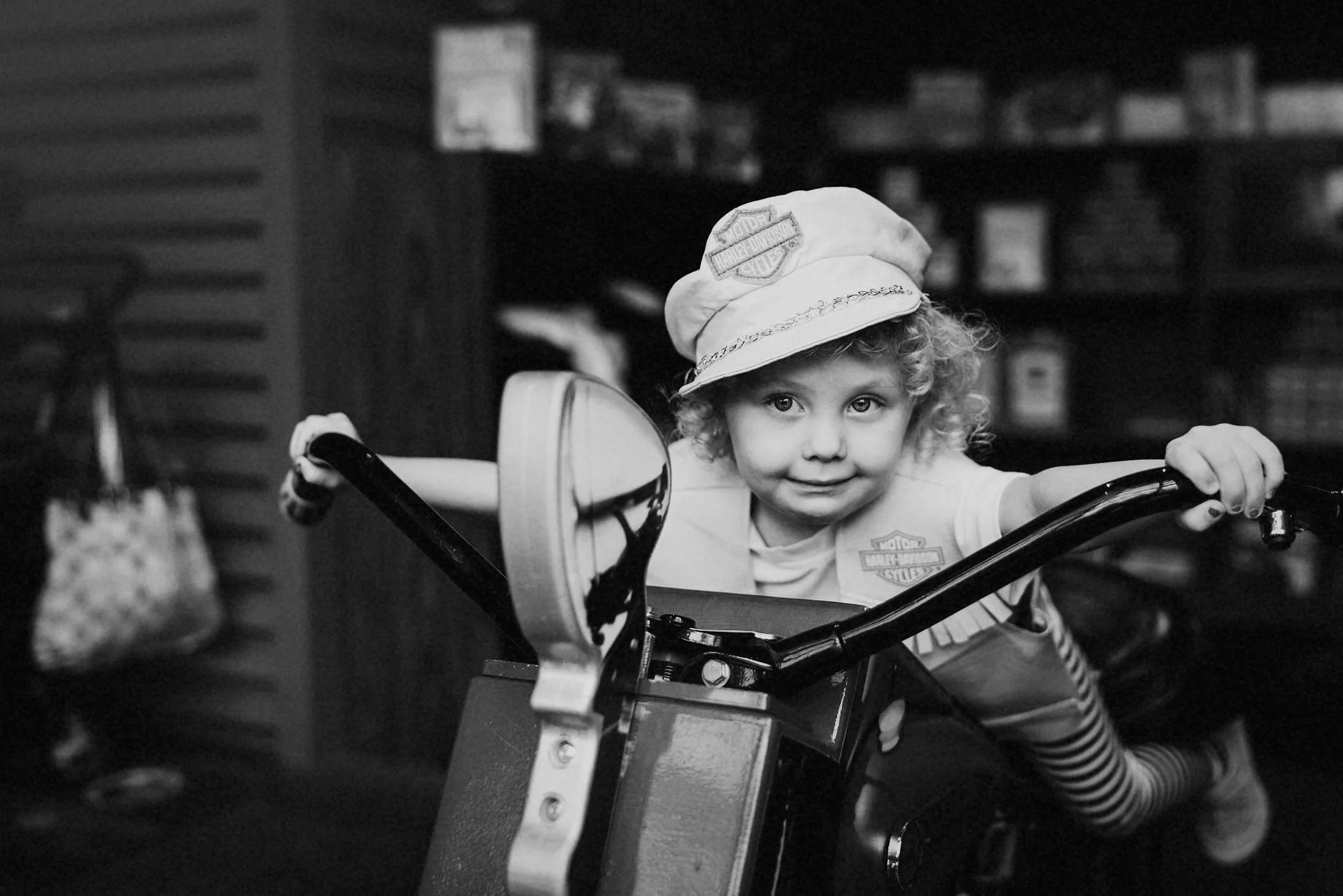 A young girl with curly hair and a cap, sitting on a child's bicycle, looking at the camera with a slight smile.