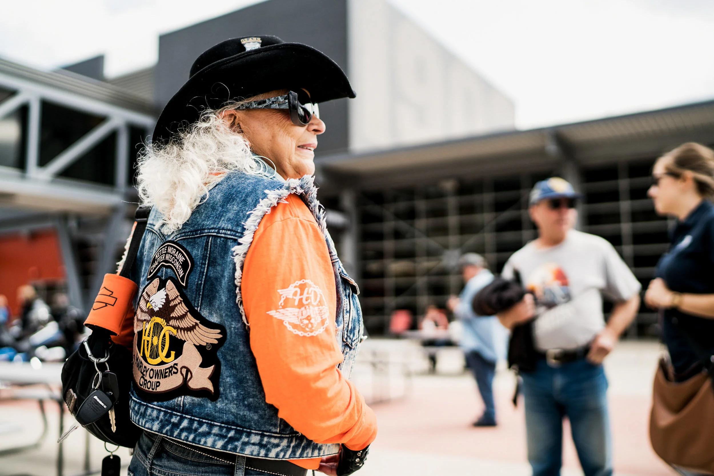 A woman with curly gray hair wearing sunglasses, a black cowboy hat, a denim vest with patches, and an orange long-sleeve shirt, standing outdoors with a group of people in the background.