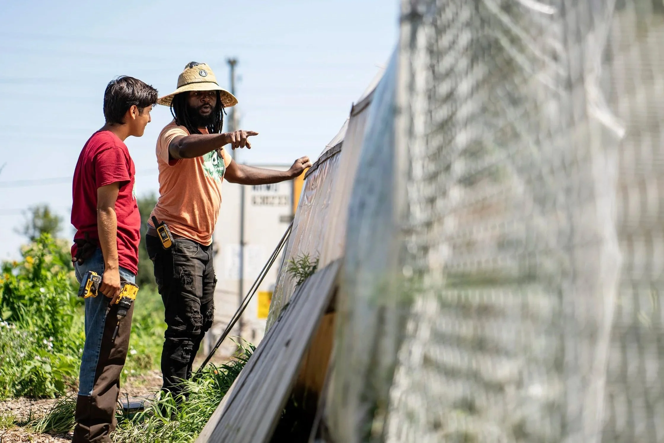 Two community farm workers installing plastic covering on a hoop house structure at an urban garden site.