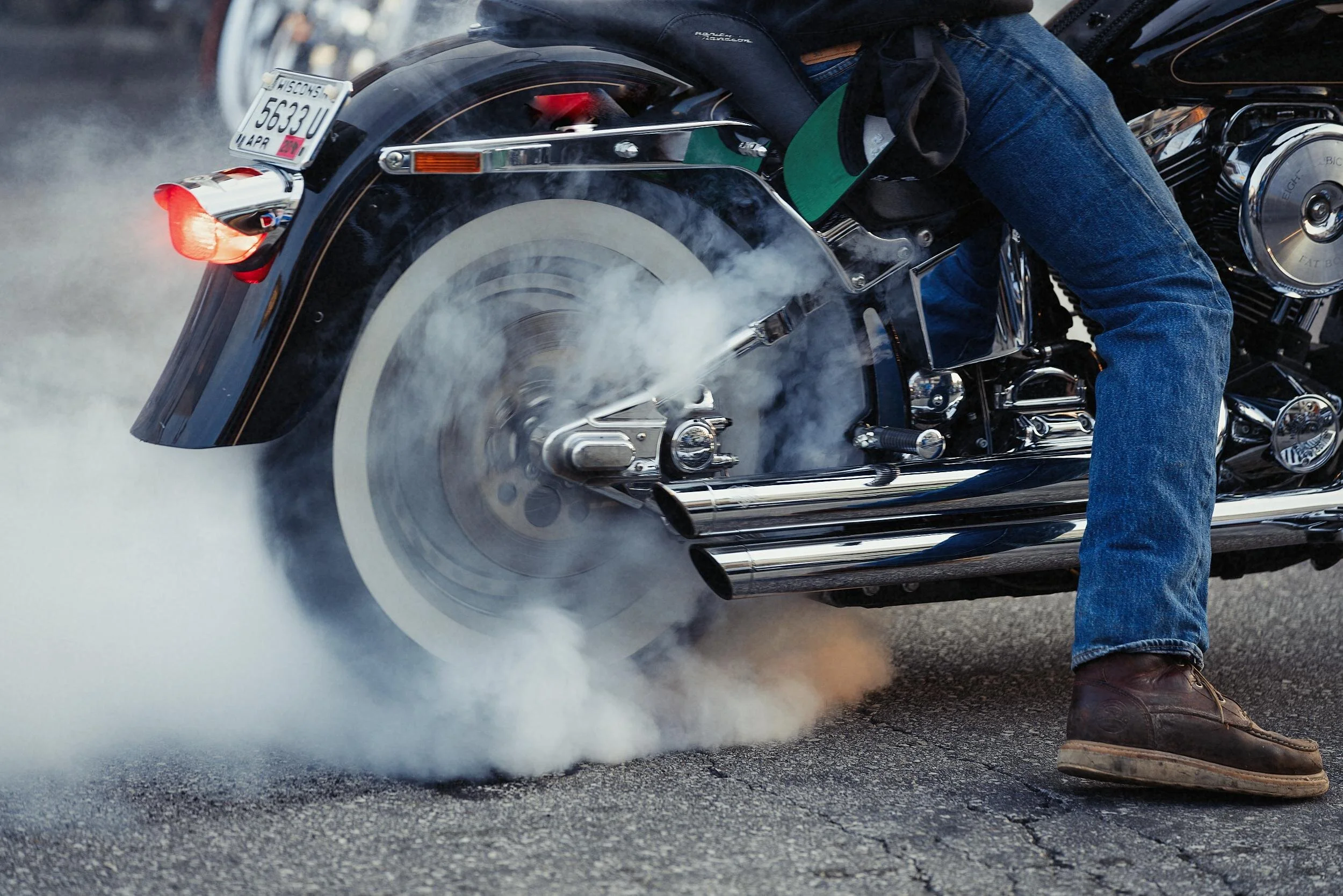Motorcycle performing burnout with smoke coming from rear tire, person wearing jeans and boots, parked on asphalt.