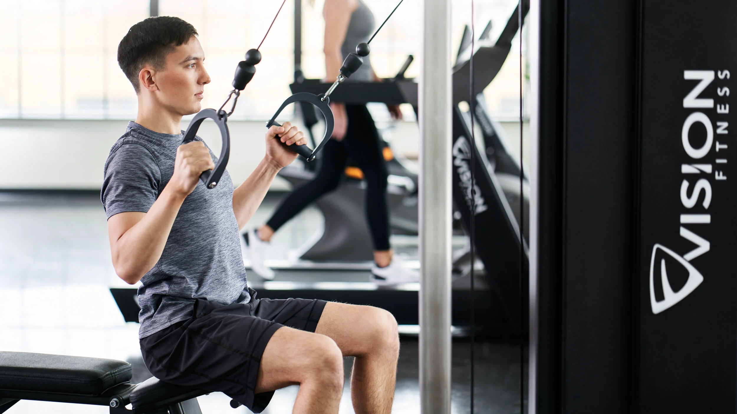 A young man performing a seated cable chest press exercise at the gym, with a woman on a treadmill in the background.