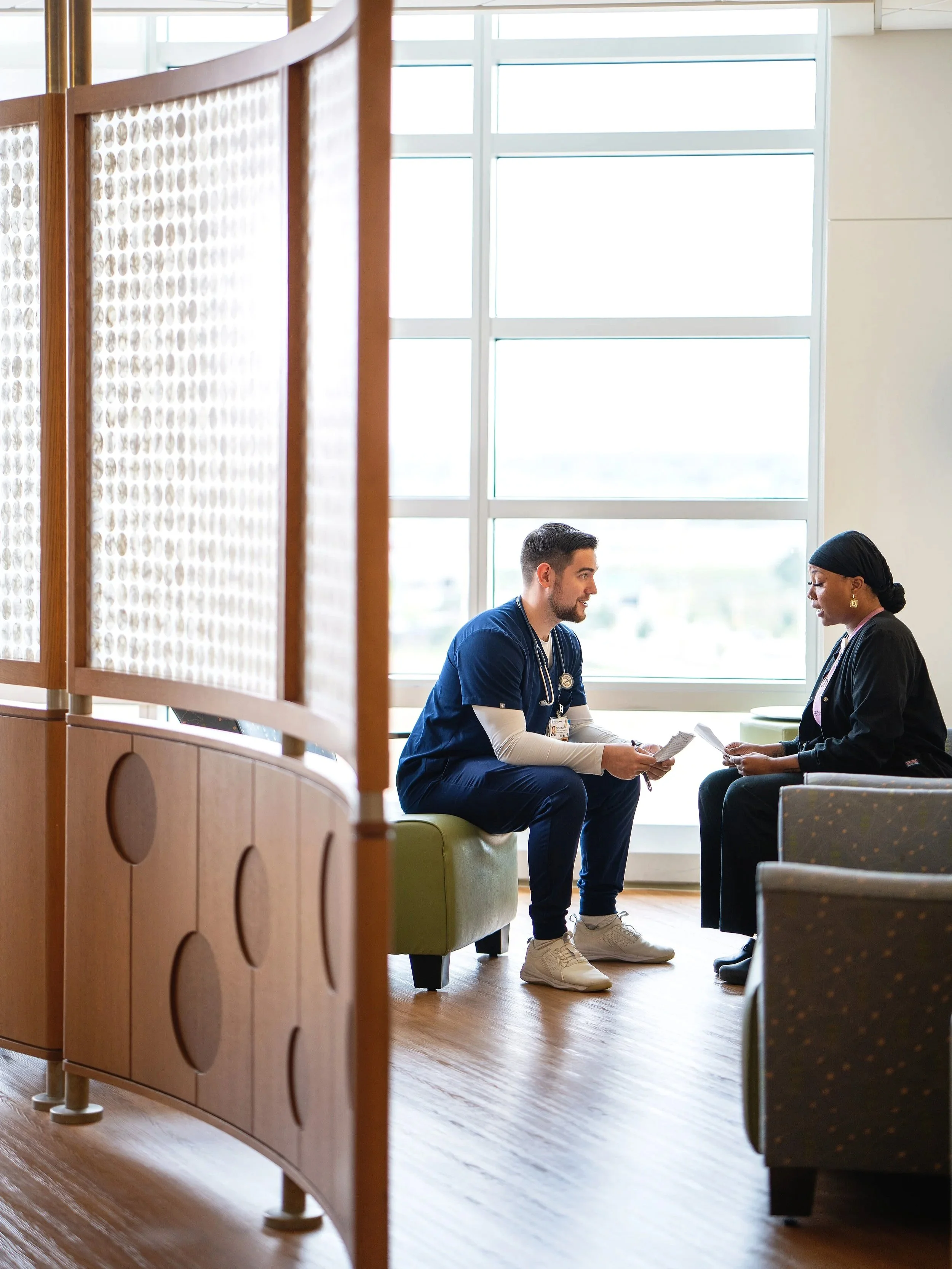 A male nurse in blue scrubs sits and talks with a Black female cancer patient in the bright waiting area of a cancer care center.